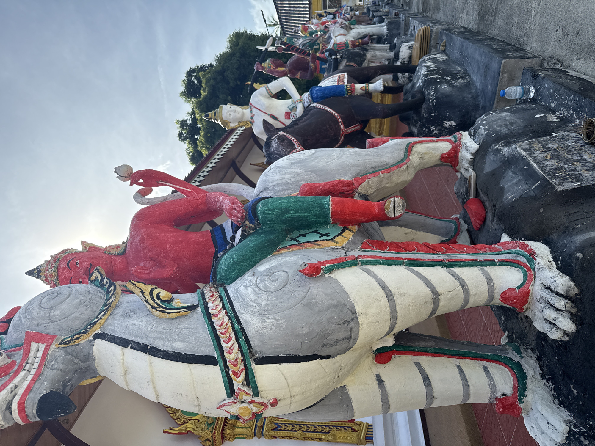 This media file shows a collection of colorful, weathered statues depicting figures on horseback, likely part of an outdoor display at a temple or religious site in Baan Lamai, Thailand. The primary subjects are the statues themselves, with a focus on a large red-skinned figure with a crown and elaborate headdress riding a white and grey horse. The horse is adorned with colorful trappings. Behind this central group, several other statues of figures on horses can be seen, some in white, brown, and black. Some figures appear to be engaged in a scene of action, suggested by the dynamic poses and the presence of weapons like spears. The statues are rendered in a folk art style with bold colors and distinct features.
The setting appears to be outdoors, with a bright sky and some greenery visible in the background. The sun is shining, suggesting it is daytime, possibly late morning or afternoon, given the angle of the light. The statues are placed on a tiered, rocky, and concrete structure. A blue plastic water bottle rests on a dark, textured surface to the right of the frame, providing a modern, mundane contrast to the ancient-feeling statues and hinting at the everyday presence of visitors or caretakers. Some of the statues show signs of age and wear, with chipped paint and some damage. The overall atmosphere is one of ancient artistry and vibrant, though perhaps slightly decaying, cultural expression. No visible text is present in the image.
