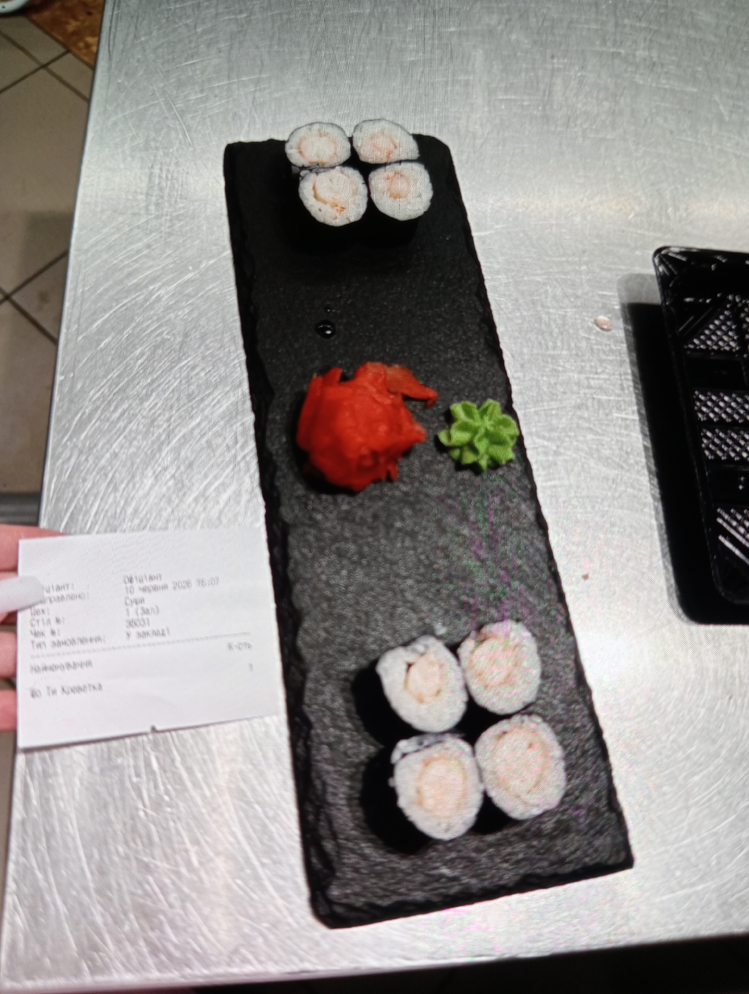 An overhead shot shows a dark slate platter with sushi, pickled ginger, and wasabi on a stainless steel surface. A receipt is held at the bottom left of the frame. The sushi appears to be shrimp rolls, with four on the top and four on the bottom of the platter. The receipt is mostly illegible but includes Cyrillic text. This scene likely takes place in Mykolaiv, Ukraine, given the Cyrillic script on the receipt, but the specific location within the city is not determinable from the image.