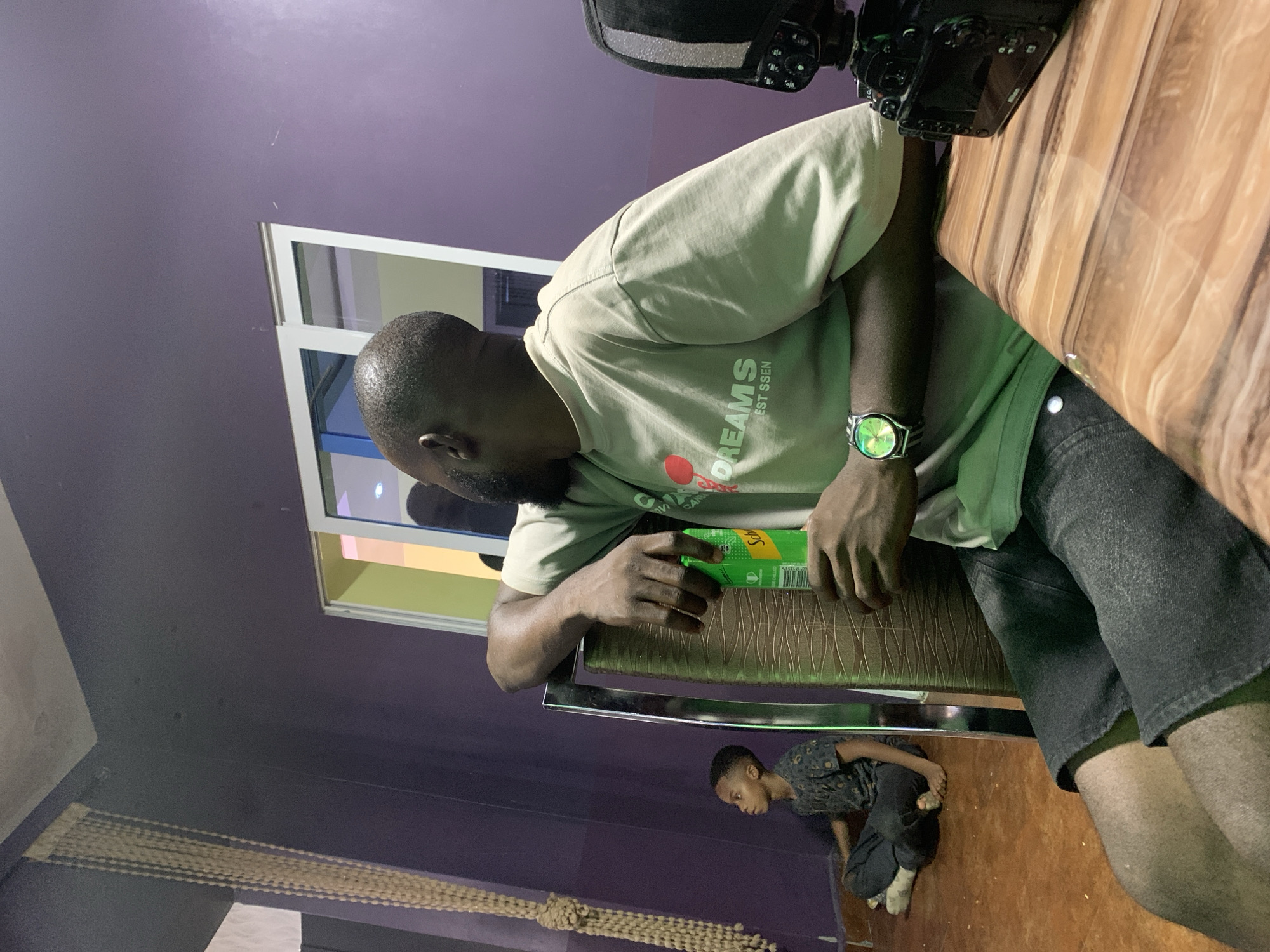 This image, reportedly located in Jos, Nigeria, depicts an indoor scene featuring two individuals and various objects within a room characterized by muted purple walls and a reddish-brown tiled floor.
In the midground, an adult male with dark skin, a short beard, and closely cropped dark hair is seated, leaning his right arm on the back of a chair that has a textured brown surface and a metallic frame. He is wearing a light beige short-sleeved t-shirt with "DREAMS" and "EST SSEN" printed in white text on the front, and dark denim shorts. A metallic wristwatch with a green face and a striped band is visible on his left wrist. He holds a green Schweppes can in his left hand, and his gaze is directed towards the viewer's left.
In the left background, a young boy, appearing to be between 5 and 10 years old, is seated cross-legged on the floor, barefoot. He wears a dark, possibly patterned, short-sleeved t-shirt and dark trousers. His attention is also directed towards the viewer's left and slightly upwards, mirroring the adult's focus.
The room's background includes a white-framed window or sliding door, reflecting ambient light and hints of light green and yellow beyond. A long, vertical decorative element, composed of light-colored beads or braided rope, hangs from the ceiling in the far left. In the right foreground, a black Nikon DSLR camera, equipped with an external flash unit, rests on a glossy table surface with a light brown and tan wood-grain pattern.