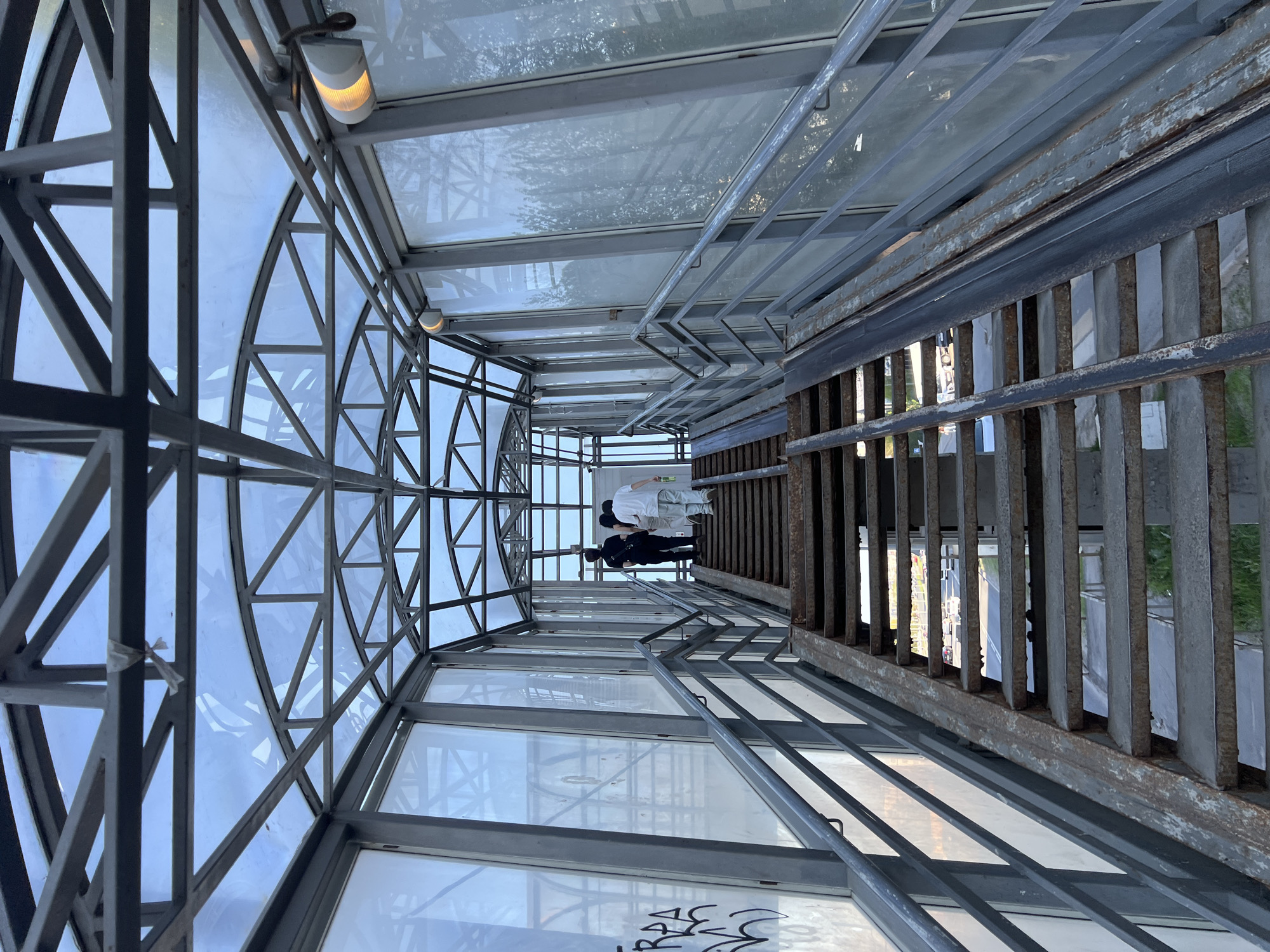 The image captures an upward-looking perspective inside a pedestrian overpass in Kazan, Russia. The overpass has a metal framework and transparent walls, offering views of the surrounding environment. Three individuals are standing towards the end of the structure, their figures partially obscured by the railings and the distance. The overall atmosphere appears to be modern and utilitarian. The scene presents a combination of architectural elements and human presence.