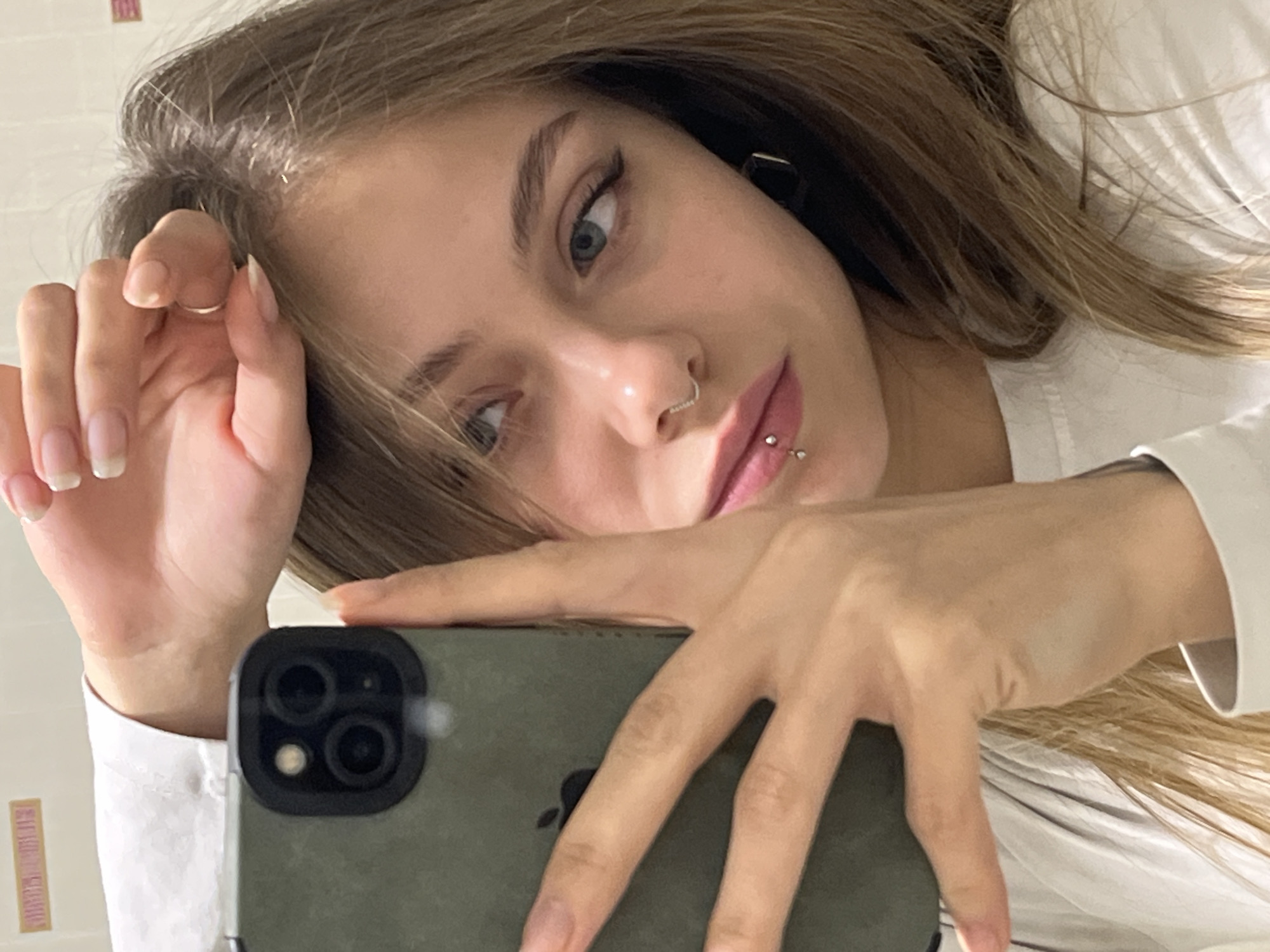A young woman is taking a selfie. She has light brown hair, light skin, and blue eyes. She is wearing a white shirt. She has piercings in her nose and lip. She is holding a smartphone in front of her face. The location is not determinable from the image.