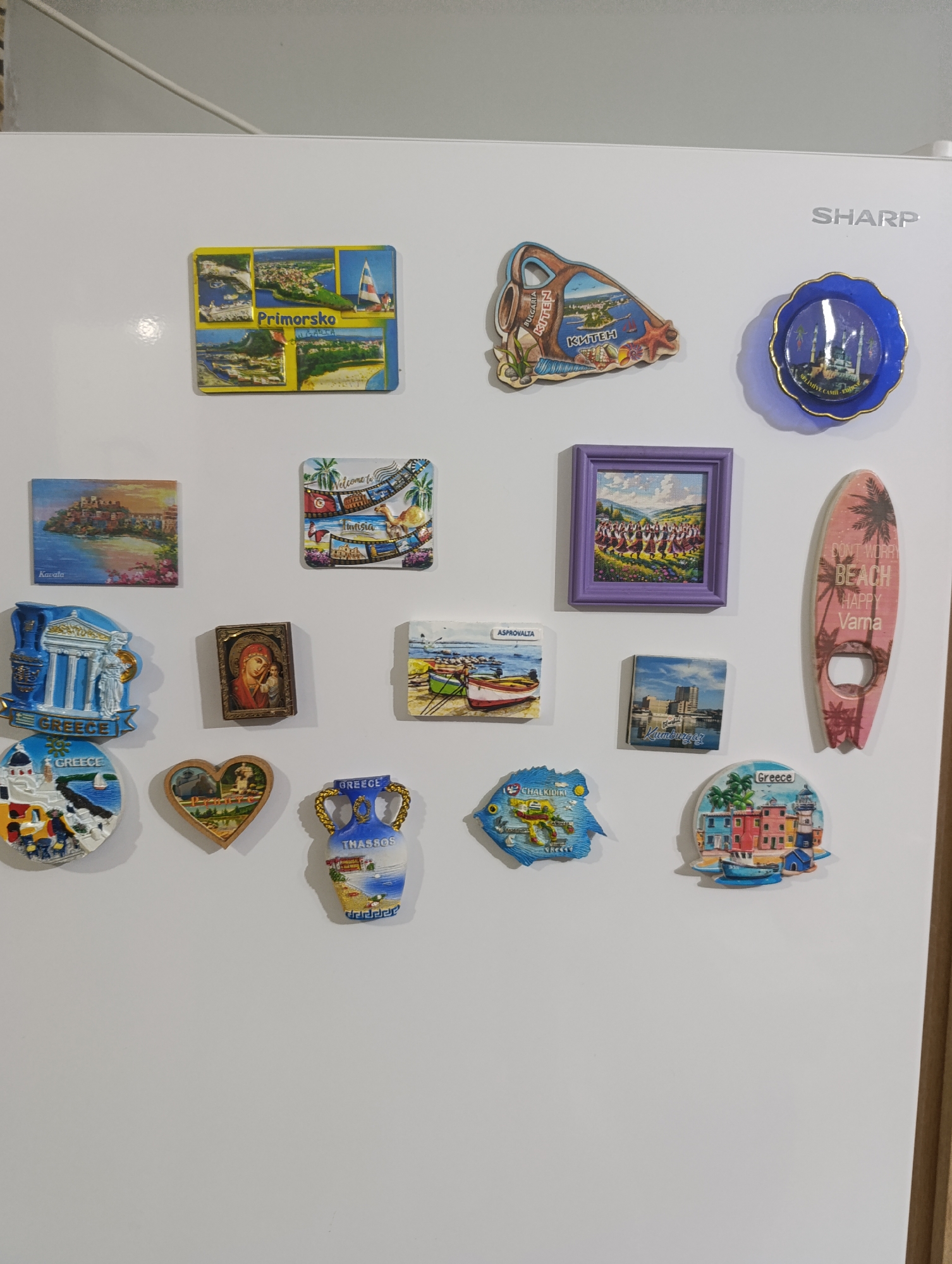 A collection of thirteen refrigerator magnets is arranged on a white refrigerator door. The magnets depict various travel destinations. From top left, clockwise, the magnets include: a four-panel magnet labeled "Primorska"; a blue decorative plate with a mosque design; a pink surfboard-shaped bottle opener with palm trees and text that reads "DON'T WORRY BEACH HAPPY Varna"; a circular magnet with a lighthouse and colorful buildings, labeled "Greece"; a fish-shaped magnet labeled "CHALKIDIKI GREECE"; a heart-shaped magnet with a landscape scene; a blue and white amphora-shaped magnet labeled "THASSOS GREECE"; a small rectangular magnet with a cityscape and text "KUMBURGOO"; a magnet depicting a small boat on water labeled "ASPROVALTA"; a framed magnet with a group of people in traditional dress; a magnet with a religious icon of the Virgin Mary and child; a square magnet with a European coastal town; and a magnet with a colorful montage of Tunisian scenes including a camel. The refrigerator door is white, and a portion of the top of a white object, possibly a cabinet, is visible at the top of the frame. The logo "SHARP" is visible on the upper right side of the refrigerator door. There are no people or animals visible in the image. The background is a plain white surface. There is no information in the image to suggest any connection to the city of Kyustendil, Bulgaria.