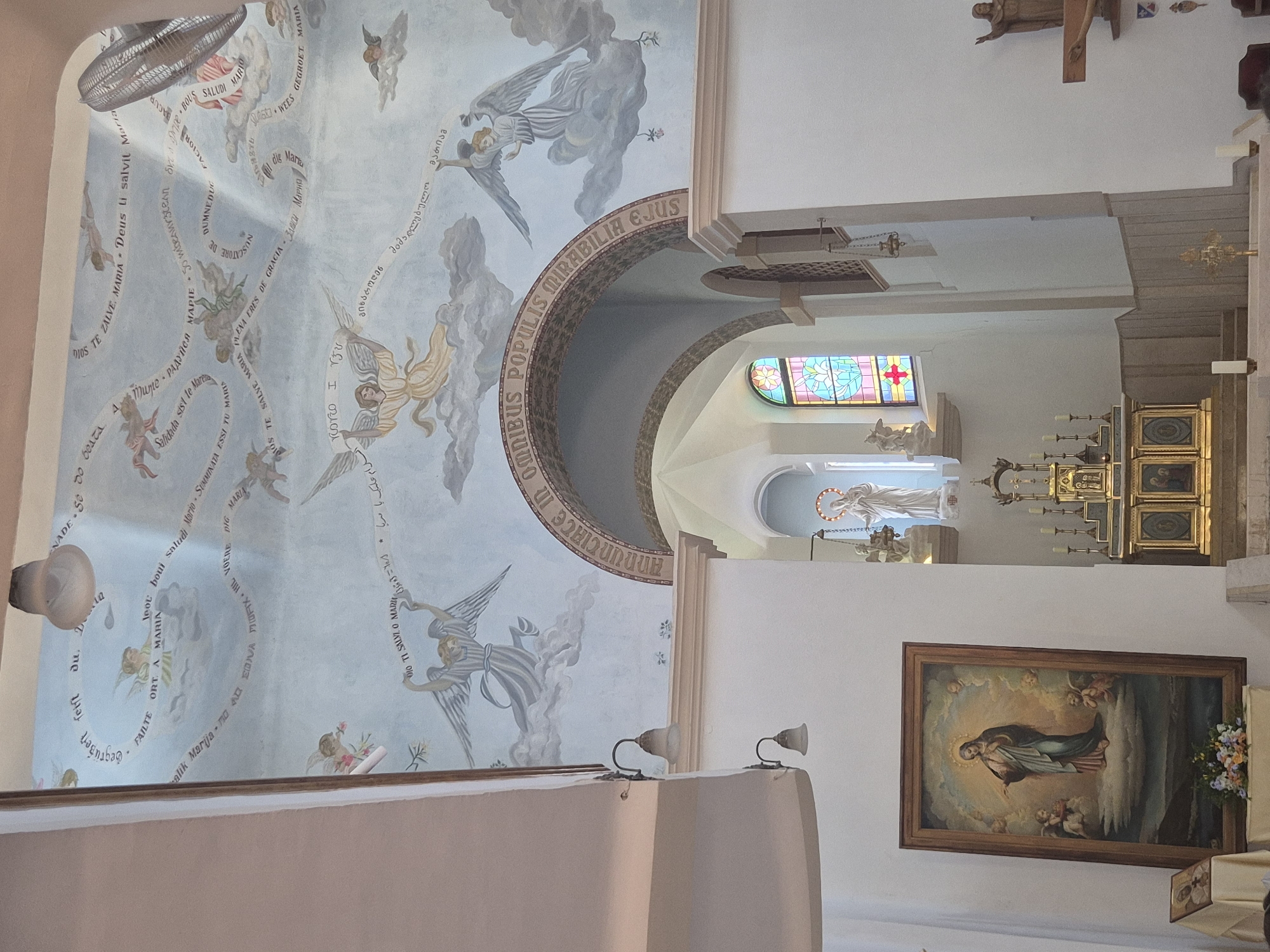 The image depicts the interior of a church, likely in the city of Mate Yehuda Regional Council, Israel, given the religious iconography and architectural style. The ceiling is adorned with a fresco featuring numerous angels in various poses, with text inscribed on banners held by some of them, suggesting a religious narrative. Directly below the fresco, an arched architectural feature is visible, with more text inscribed around its curve.
In the foreground, to the left, is a large framed painting of the Virgin Mary surrounded by angels. A smaller framed image and a floral arrangement are placed in front of it. Towards the center and right of the image, an altar is visible, adorned with candles and decorative elements. A statue of a religious figure is positioned on a raised platform behind the altar. Further to the right, a wooden crucifix is mounted on the wall. The environment appears to be well-lit, with natural light entering from a stained-glass window above the altar. The overall scene suggests a place of worship or religious significance.