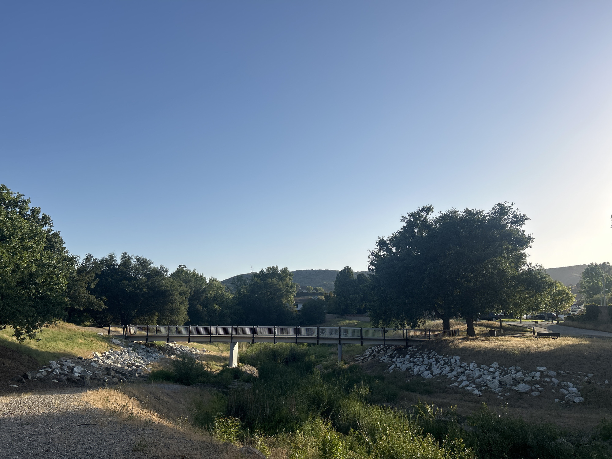 The image displays a serene outdoor landscape, characteristic of a park or natural area in Agoura Hills, United States of America. A pedestrian bridge, featuring a wooden deck and dark metal railings, serves as a central subject, spanning across what appears to be a dry creek bed or seasonal stream. The banks of this waterway are fortified with numerous large, light-colored rocks.
The scene is rich with lush, mature trees, possibly oaks, boasting dense green canopies. Several prominent trees frame the foreground on both the left and right, while more abundant foliage and verdant hills rise in the midground and background. The sky overhead is a clear, bright blue, with a noticeable brightness gradient on the right side, indicating the sun is low on the horizon. This lighting suggests either early morning or late afternoon, as warm sunlight illuminates patches of the dry, golden-brown grass and the upper parts of the trees, creating areas of light and shadow.
A gravel path is visible in the lower-left corner, hinting at walking trails. The overall setting evokes a sense of peaceful natural beauty. No people are visible within the frame, and there is no discernible text present in the image.