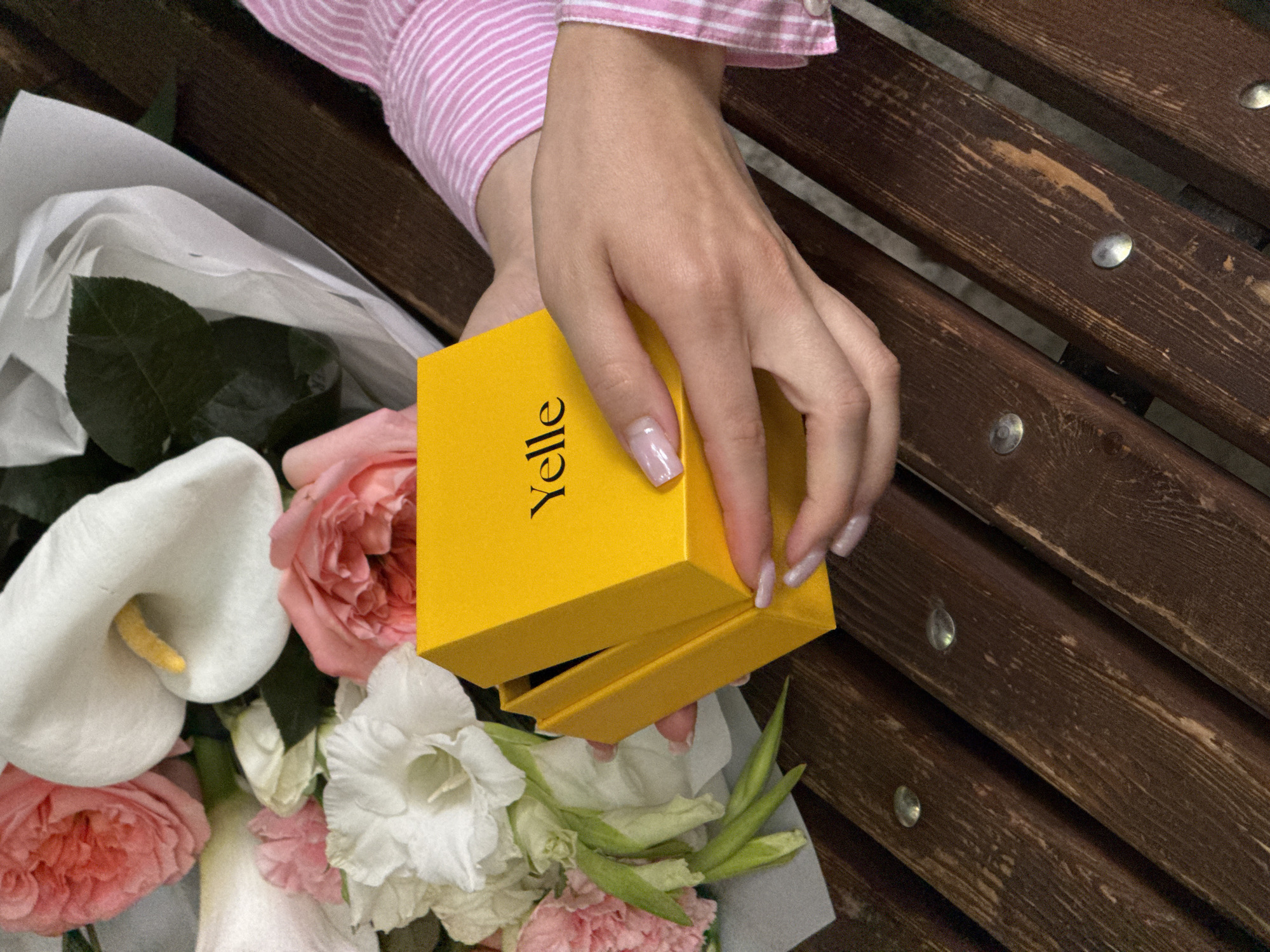 A close-up, high-angle shot shows a person's hand opening a small, bright yellow gift box. The hand, with light skin tone and manicured nails painted in a light, shimmery pink or lavender shade, holds the box open, revealing its interior. The word "YELLE" is prominently displayed vertically in black text on the side of the yellow box.
To the left and below the hand and box, a lush bouquet of flowers is visible, wrapped in white paper. The bouquet includes several soft pink roses, a distinctive white calla lily with a yellow spadix, and white gladioli-like flowers, interspersed with green foliage.
The person's arm, dressed in a pink and white striped long-sleeved shirt or blouse, is visible in the upper left corner. The setting appears to be an outdoor wooden bench, with dark brown, slightly worn wooden slats and silver circular rivets visible in the background, primarily on the right side of the image. The lighting is bright, suggesting daytime. The overall scene conveys a moment of unwrapping a gift, possibly in a park or garden setting.