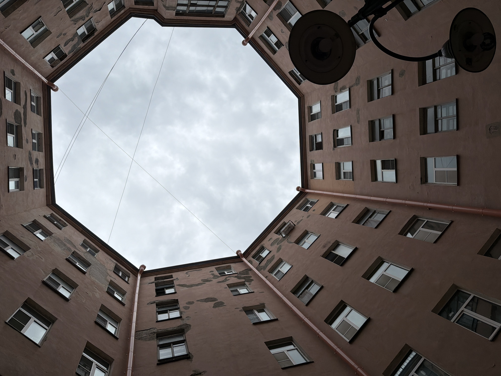 This is a low-angle, upward shot of a St. Petersburg courtyard, also known as a "well courtyard." The buildings surrounding the courtyard are multi-story, with numerous windows visible on their facades. The buildings are painted a muted reddish-brown color, and some areas show signs of wear and peeling paint. Red pipes run vertically along the exterior walls, likely for drainage. The sky above is overcast and gray, with visible power lines crisscrossing the open space. A street lamp with two light fixtures is partially visible in the foreground, obscuring some of the building details. The architecture suggests a typical residential building in the historic center of St. Petersburg. There are no people visible in the frame.