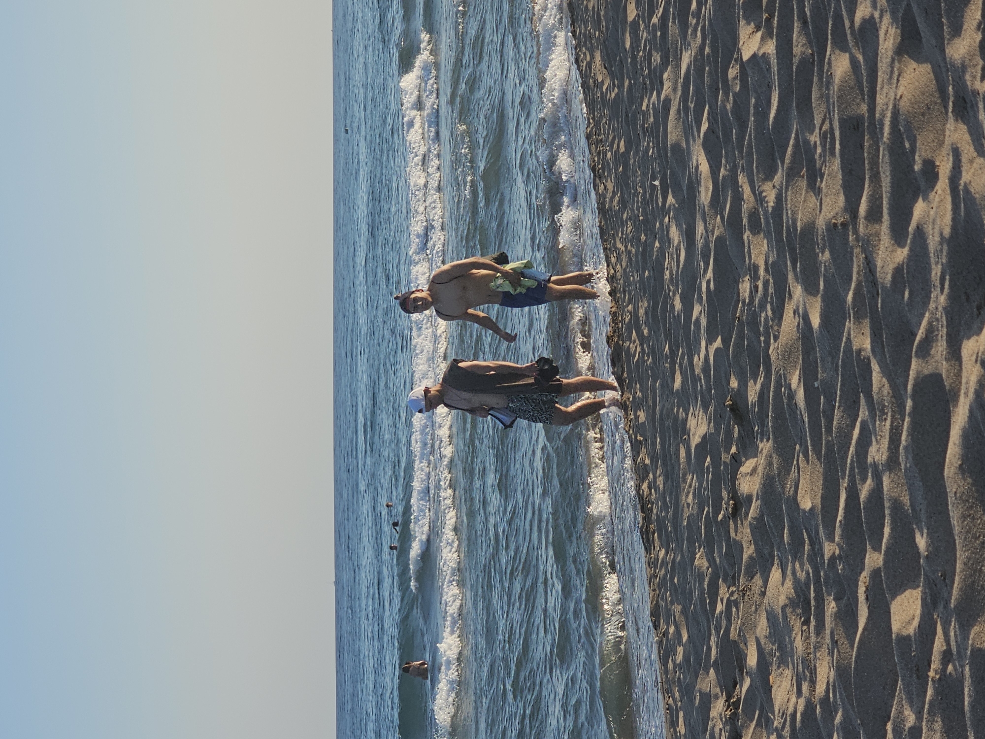 The image, taken at Castelldefels, Spain, shows a beach scene with two people visible. The perspective is from a high angle, looking down at the shoreline. The right side of the frame is dominated by textured sand, likely from the dunes of Castelldefels beach, with distinct shadows indicating a low sun angle, suggesting either early morning or late afternoon.
To the left, the shallow, blue water of the Mediterranean Sea is visible, with small waves breaking near the shore. The sky is a pale blue, transitioning to a soft white near the horizon, indicating clear weather.
In the water, near the left edge of the frame and partially obscured by waves, is a person. Closer to the shore, standing at the water's edge with their feet in the shallow water, is another person. This individual is wearing swim trunks and appears to be holding a light-colored towel or piece of fabric. Their posture is relaxed. Further out in the water, several small, dark objects can be seen, possibly buoys or other beachgoers.
The overall mood of the image is peaceful and leisurely, capturing a moment of relaxation by the sea. There is no visible text in the image.