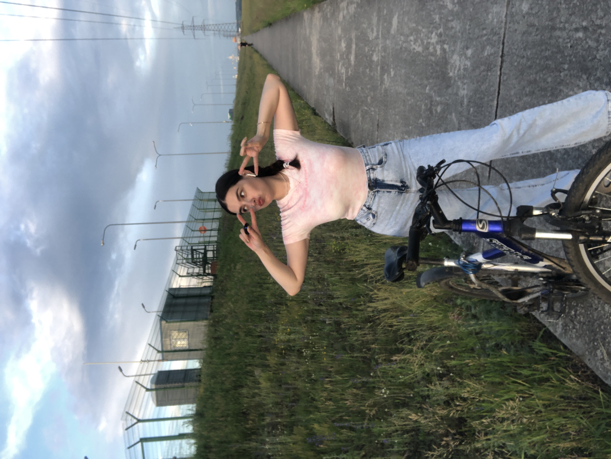 A young woman with dark hair in braids and wearing a pink tie-dye t-shirt and light-wash jeans stands on a paved path next to a bicycle. She is posing with peace signs near her face and wearing white earbuds. Behind her, a grassy embankment slopes upwards, topped with a chain-link fence and some small industrial buildings. The path extends into the distance, lined with streetlights and leading towards power lines and a hazy horizon. The sky is overcast with dramatic clouds. The scene appears to be in an outdoor, possibly industrial or semi-urban, location in Noviy, Russia.