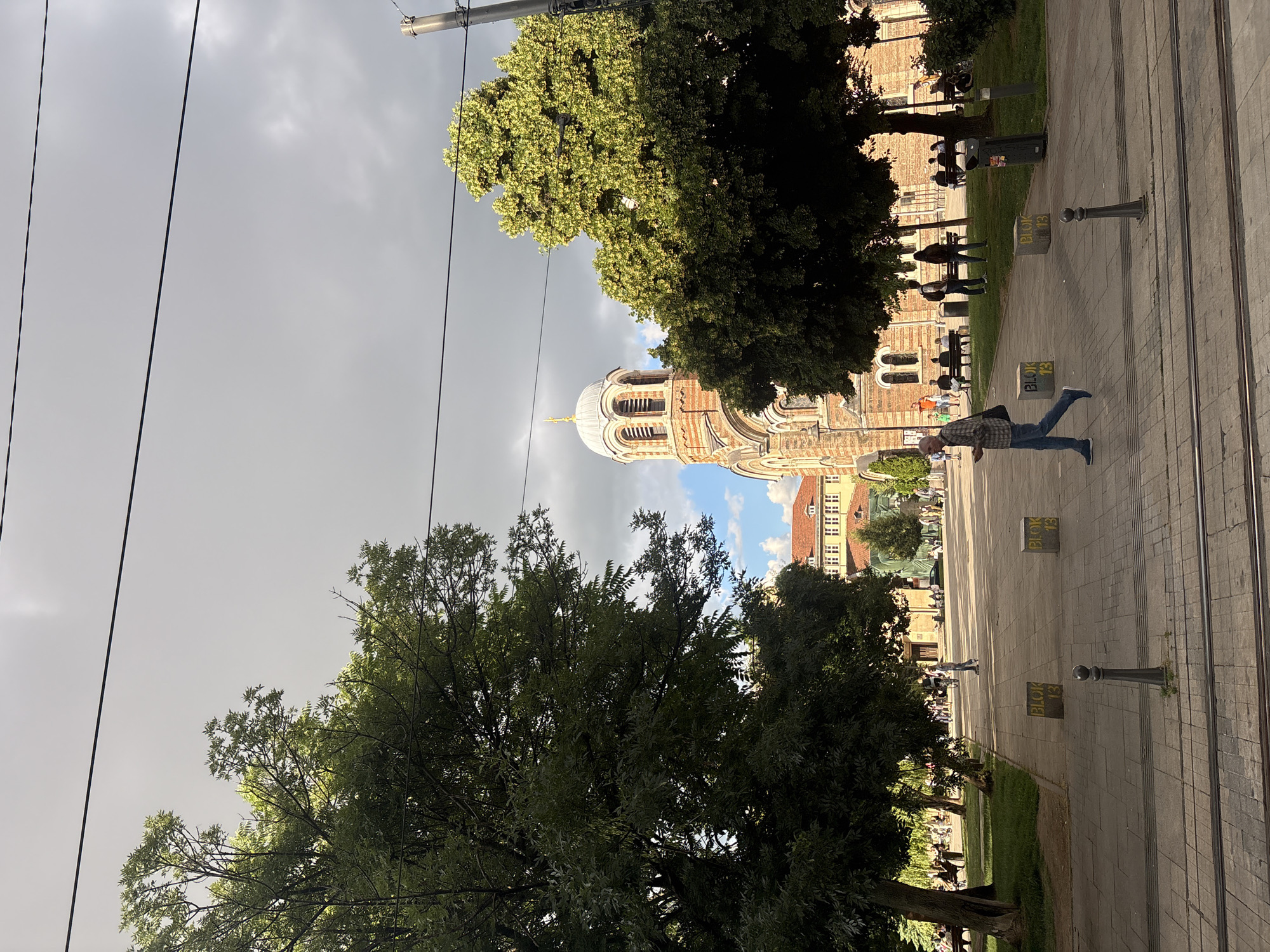 The image depicts an outdoor urban scene featuring a large park area with mature trees on the left and right, and a church with a prominent dome and brick facade in the background. A paved walkway extends through the park, with several people walking and sitting on benches. A man in a plaid shirt and blue jeans is walking across the walkway in the foreground. Two concrete blocks with yellow lettering "BLOK 13" are visible on the walkway. The sky is partly cloudy with areas of both bright sunlight and dark clouds. Overhead power lines are visible.