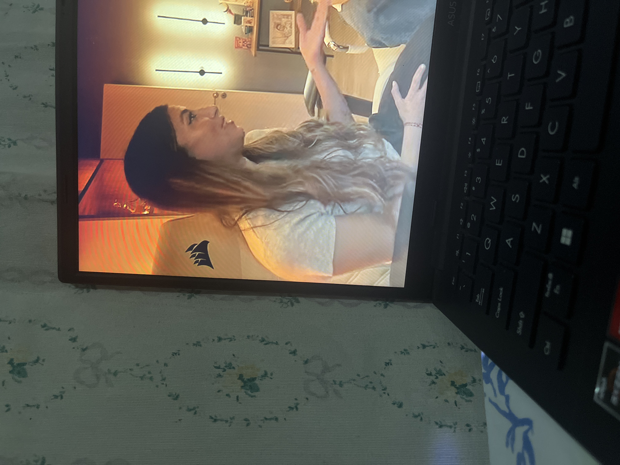 The image shows a laptop screen displaying a woman with long, wavy brown hair wearing a light-colored t-shirt. She is seated and appears to be talking, with her right hand gesturing outwards. Behind her, on the screen, is a room with warm lighting and decorative elements on a shelf. The laptop itself is black and shows a portion of its keyboard. The background of the photo is a light-colored wall with a floral pattern. The scene is captured indoors, likely in a residential setting. The laptop is positioned on a surface with blue and white patterned fabric. There is no identifiable information about the specific location within Santa Ana, Venezuela.