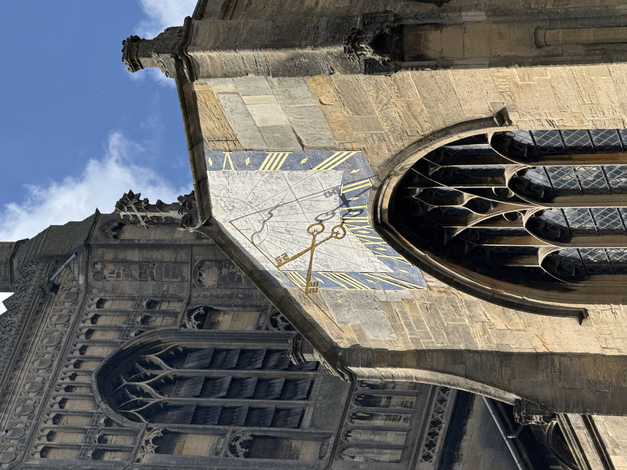 This is a low-angle shot of a stone building in Norwich, United Kingdom. The building is adorned with ornate carvings and features. A large sundial is mounted on the side of the building, with Roman numerals indicating the hours. The sundial's gnomon casts a shadow, suggesting the time of day. The building also has stained-glass windows. The sky above is blue with some clouds.