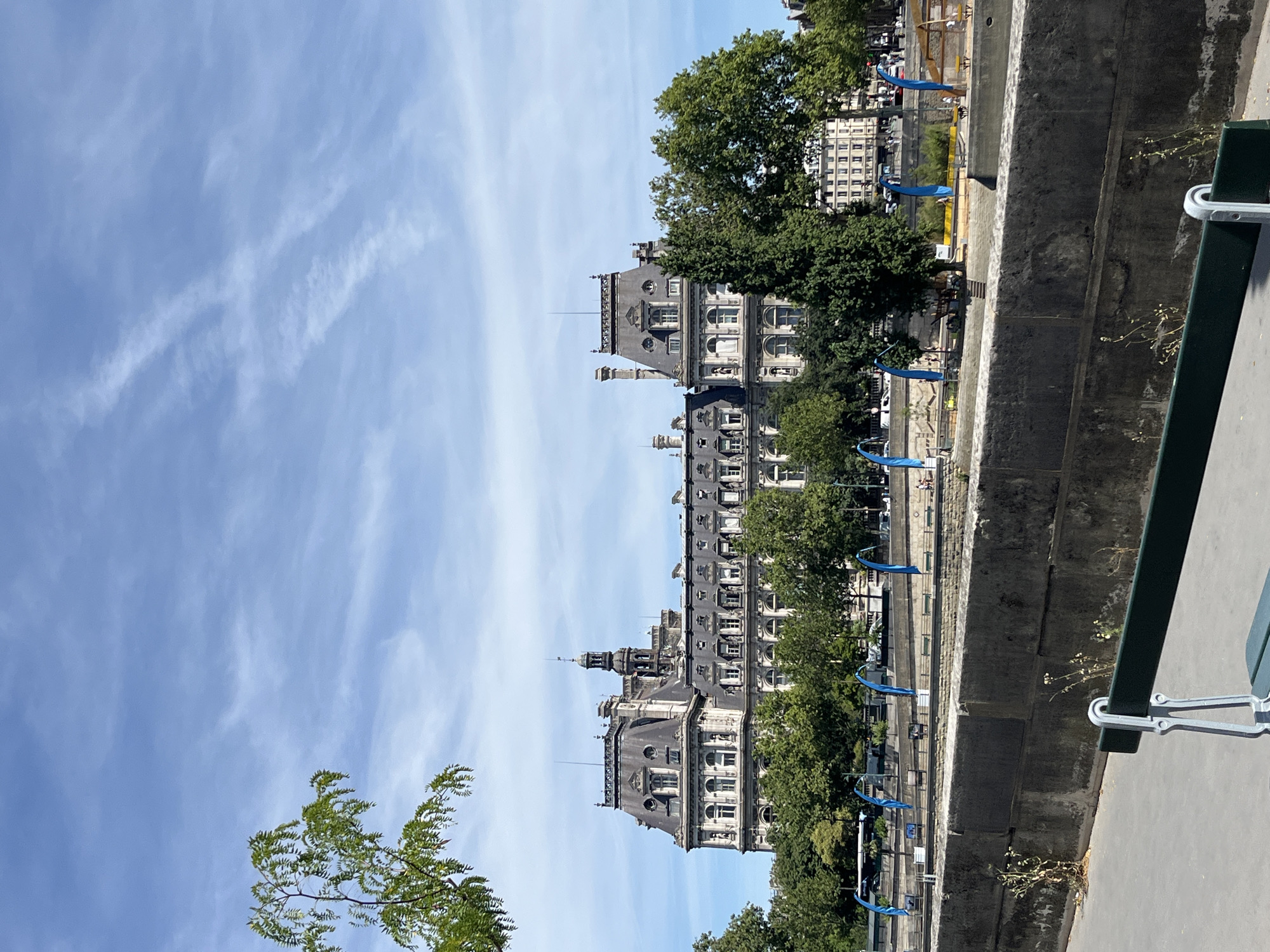 In Paris, France, a grand historic building with ornate architecture and a mansard roof stands against a bright blue sky with wispy clouds. The building, which appears to be the Hôtel de Ville, is framed by lush green trees on its lower levels. In the foreground, a concrete embankment borders a body of water. Beyond the embankment, a pathway is visible with a dark green bench in the lower right corner. Blue flags are planted along the riverbank further in the distance, and there are some people walking and sitting in the area.