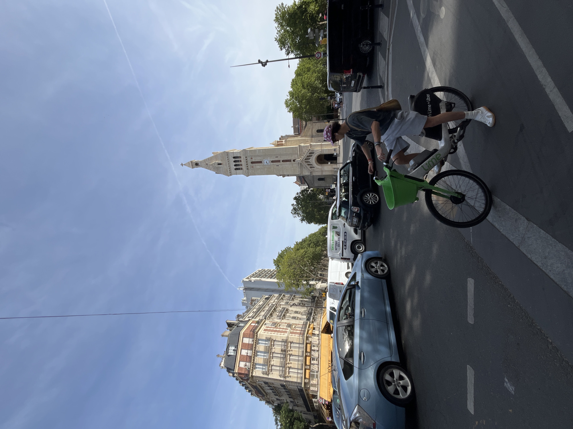 The image shows a street scene in Paris, France, dominated by a tall church with a clock tower. In the foreground, a person is riding a shared electric bicycle with a green basket. The person is wearing a t-shirt, shorts, a hat, and white sneakers. They are carrying a brown bag over their shoulder. The bicycle has "Lime + Uber" branding on the back. Several vehicles are visible in the background, including a light blue hybrid car, a black mini-car, a white van, and a black van. The buildings in the background are typical Parisian architecture, with multiple stories and balconies. Trees line the street, and the sky is blue with some faint clouds and a contrail. The scene appears to be a sunny day.