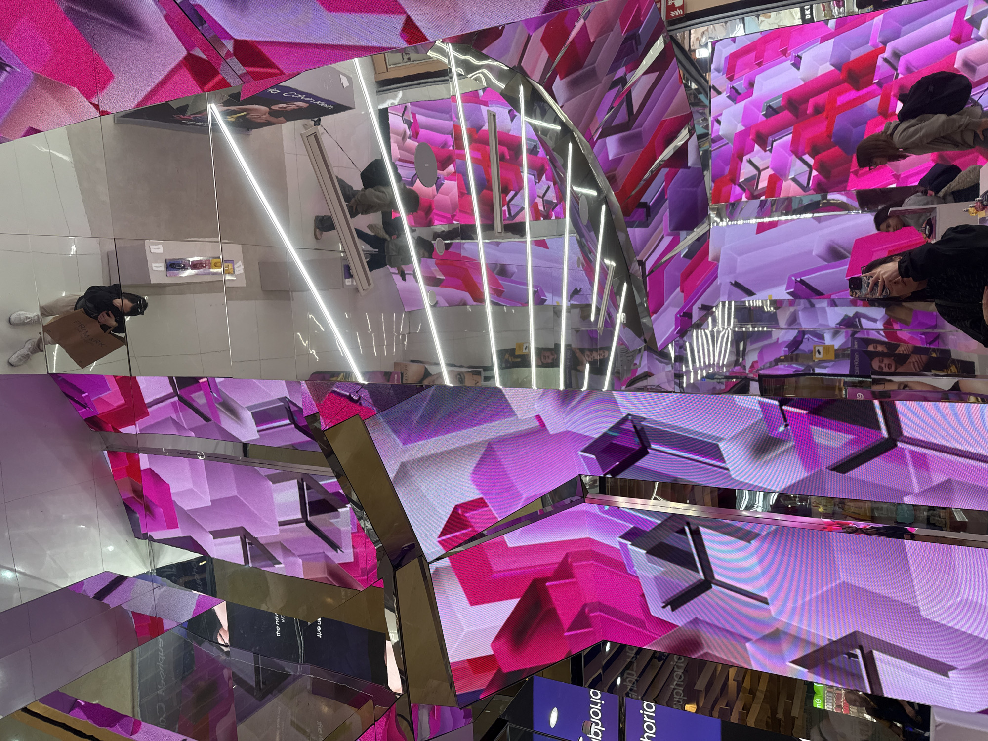 This image captures a dynamic indoor commercial environment, likely a modern shopping mall or retail store, located in Barcelona, Spain. The scene is dominated by an architectural installation featuring extensive mirrored surfaces and large digital screens, creating a complex, kaleidoscopic effect through reflections.
The screens display vibrant, abstract geometric patterns of 3D blocks in shades of magenta and purple, giving the space a futuristic and energetic aesthetic. Numerous linear white lights, possibly LED strips, are reflected repeatedly, further enhancing the visual complexity.
Several individuals are visible within the reflections. In the bottom left, a person wearing a black top and light-colored pants holds a "PRIMARK" shopping bag, indicating a shopping activity. Another person towards the right is seen holding a smartphone. Other blurry figures are scattered throughout the reflections, suggesting a bustling environment.
Visible text includes "PRIMARK" on a shopping bag, and reflected advertisements like "Calvin Klein" and "euphoria" (likely referring to the Calvin Klein fragrance). Other partial texts such as "Azorique," "the nev," "an erit," and "time" are also decipherable from the reflections. The floor appears to be a light, polished surface. The overall atmosphere is modern, bright, and visually stimulating due to the interplay of light, digital displays, and reflections.