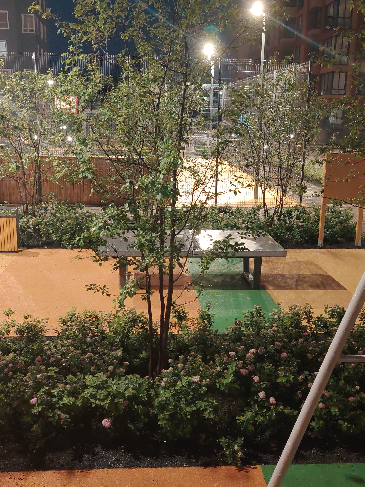 In an urban courtyard at night, a concrete ping-pong table stands on a multi-colored paved surface, a blend of orange and green. The area is bordered by lush green bushes with small pink flowers, and several trees with sparse leaves. In the background, a chain-link fence encloses a court, illuminated by streetlights that cast a yellow glow. Apartment buildings with lit windows rise on either side of the courtyard, indicating a residential area. The overall scene suggests an outdoor recreational space in a city.