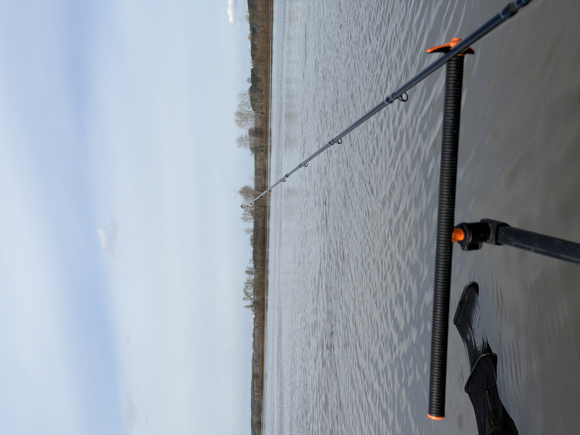 The image, when properly oriented, shows a wide body of water, likely a river, under an overcast sky. A fishing rod, light grey with several line guides, extends from the lower right foreground towards the center-left horizon. The rod is resting on a black, ribbed rod holder or bank stick, which is partially submerged in the water on the right side of the frame. Below the rod holder, a black boot or shoe is also partially submerged in the water.
The water surface is textured with small ripples. Across the mid-ground, a distant bank lined with bare trees stretches horizontally, indicating a wide expanse of water. The sky above is mostly overcast, with patches of lighter grey and soft blue. The overall impression is one of a calm, perhaps chilly, daytime scene, possibly in early spring or late autumn given the lack of foliage on the trees. The activity taking place is fishing. No visible text is present in the image. The location is Nikolo-Berezovka, Russia.