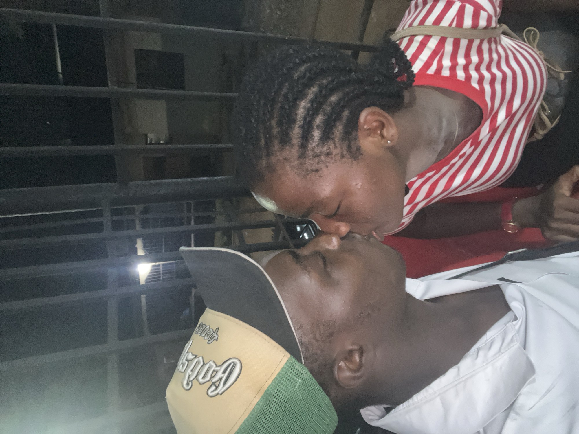 The image shows a close-up, intimate shot of a young African couple kissing. The man, on the left, is wearing a tan and green trucker hat with "Gods Plan" embroidered on it, and a white collared shirt. He has his eyes closed. The woman, on the right, has her eyes closed and her hair styled in cornrows. She is wearing a red and white striped t-shirt. They are positioned in front of a dark metal fence or gate, with a dimly lit building visible in the background. The setting appears to be outdoors at night, possibly in the city of Nkpor, Nigeria, given the context.