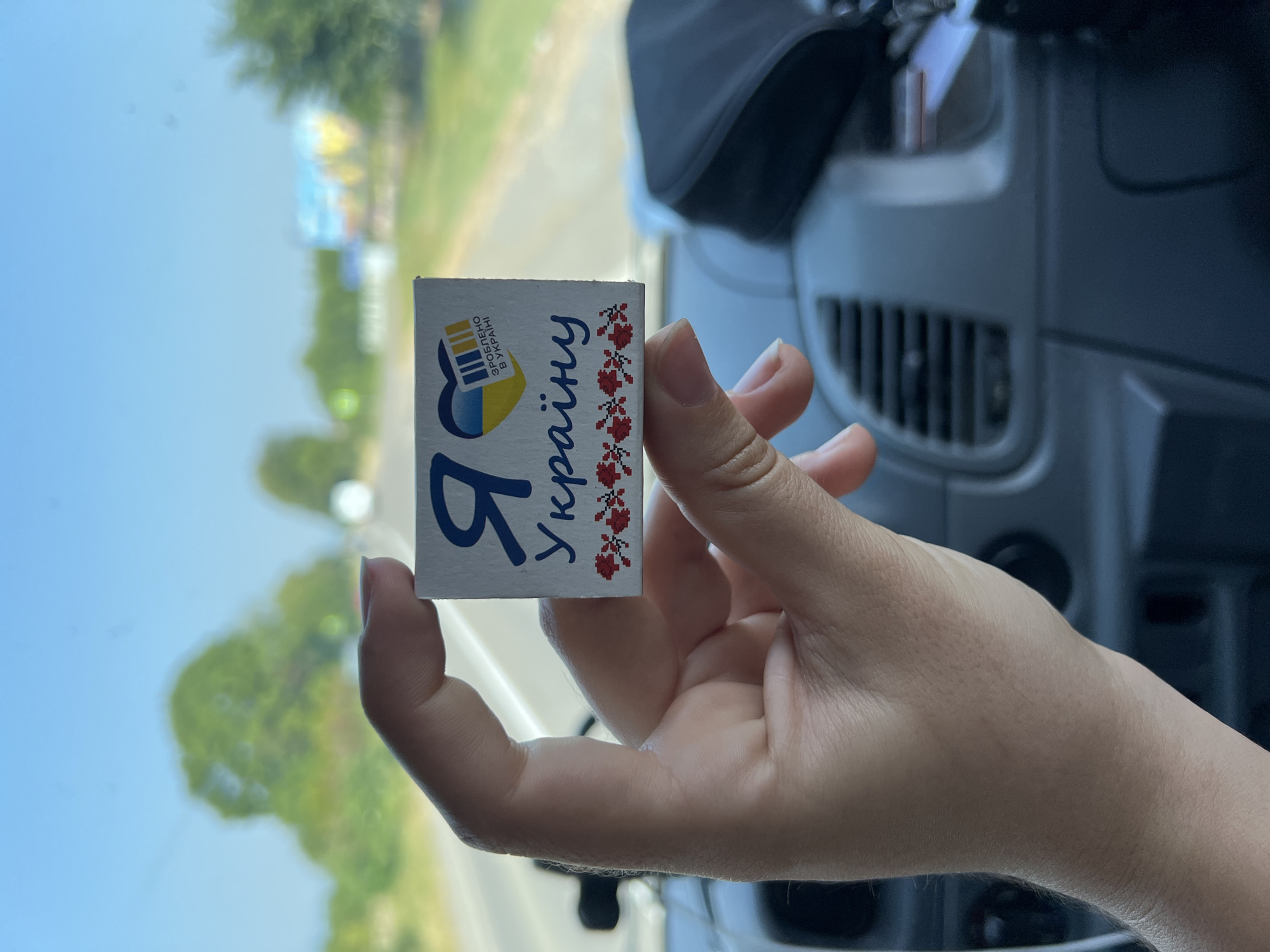 A person's hand holds a small, square item with text and a heart design against a blurred background of a road and trees. The item is white with blue and yellow elements, suggesting a connection to Ukraine. The background appears to be a sunny day with a clear blue sky, consistent with an outdoor setting likely in a rural area or on the outskirts of a city. The overall image is a close-up of the item being held, with the environment serving as a backdrop. The specific location of Ruske Pole, Ukraine, is not directly discernible from the visual elements present.