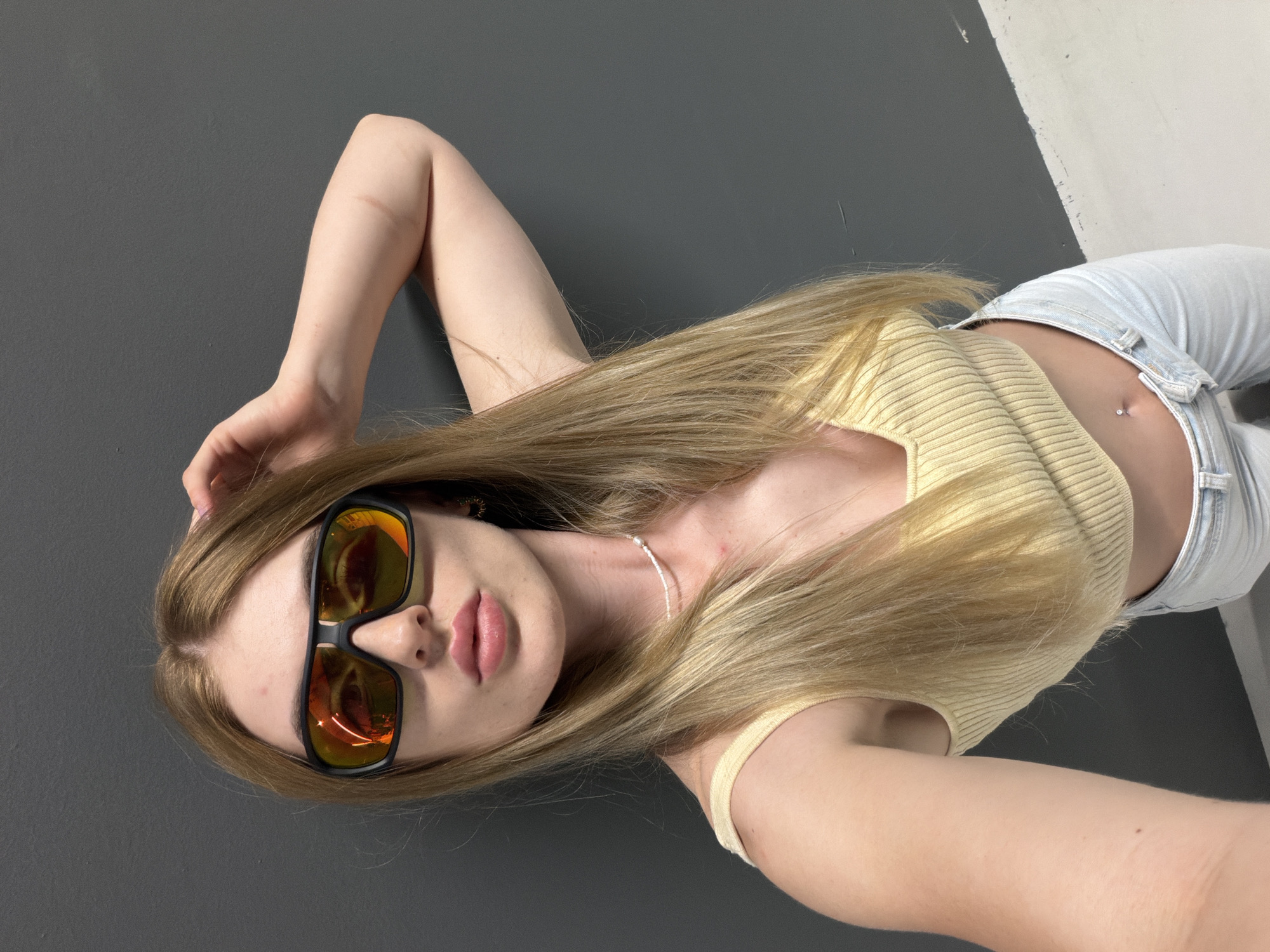 A young woman with long blonde hair and an orange-tinted aviator-style sunglasses poses for a selfie against a dark gray wall. She is wearing a light yellow ribbed crop top and light blue jeans. Her right hand is raised, touching her hair. A subtle pearl necklace is visible around her neck. She has a navel piercing with a small silver stud. The bottom portion of the image shows a white floor that transitions to the dark gray wall. The scene appears to be indoors, possibly a studio or a plain-colored room.