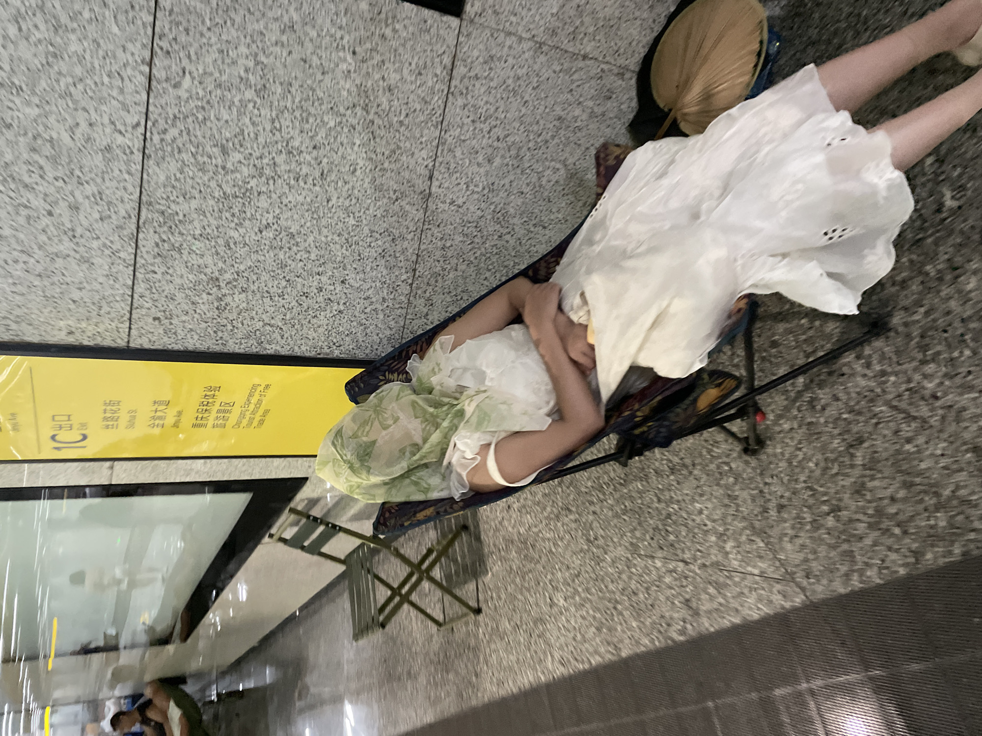 In a modern urban environment, likely a subway station in Liangjiang, China, a person is resting in a portable chair, their face obscured by a sheer, green patterned scarf. They are wearing a flowing white dress. Next to the chair, a folded green metal chair is visible. In the background, a yellow sign with Chinese characters and English text reads "1C Exit" and lists street names such as "Sizhua St." and "Jinyu Ave." Behind the resting person, a wall composed of speckled gray granite tiles is evident. A large glass partition runs along the left side of the image, reflecting the surrounding environment and other individuals who appear to be waiting or resting. The floor is a speckled, tiled surface. A traditional-looking fan lies on the ground next to the chair.