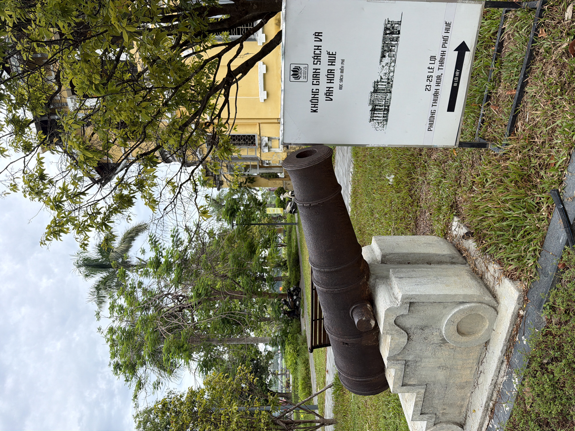 In the foreground of the image, an old, weathered cannon rests on a concrete pedestal in a grassy park. To the right of the cannon, a white sign with black lettering provides information about a location, including an address: "23-25 Le Loi, Thuan Hoa Ward, Hue City." The sign also features a sketch of a building and an arrow pointing to the right, indicating direction. In the background, a large, yellow building with traditional architecture stands behind lush green trees. The sky is overcast. The scene appears to be an outdoor public space, possibly a historical site or museum exhibit, situated in a city.