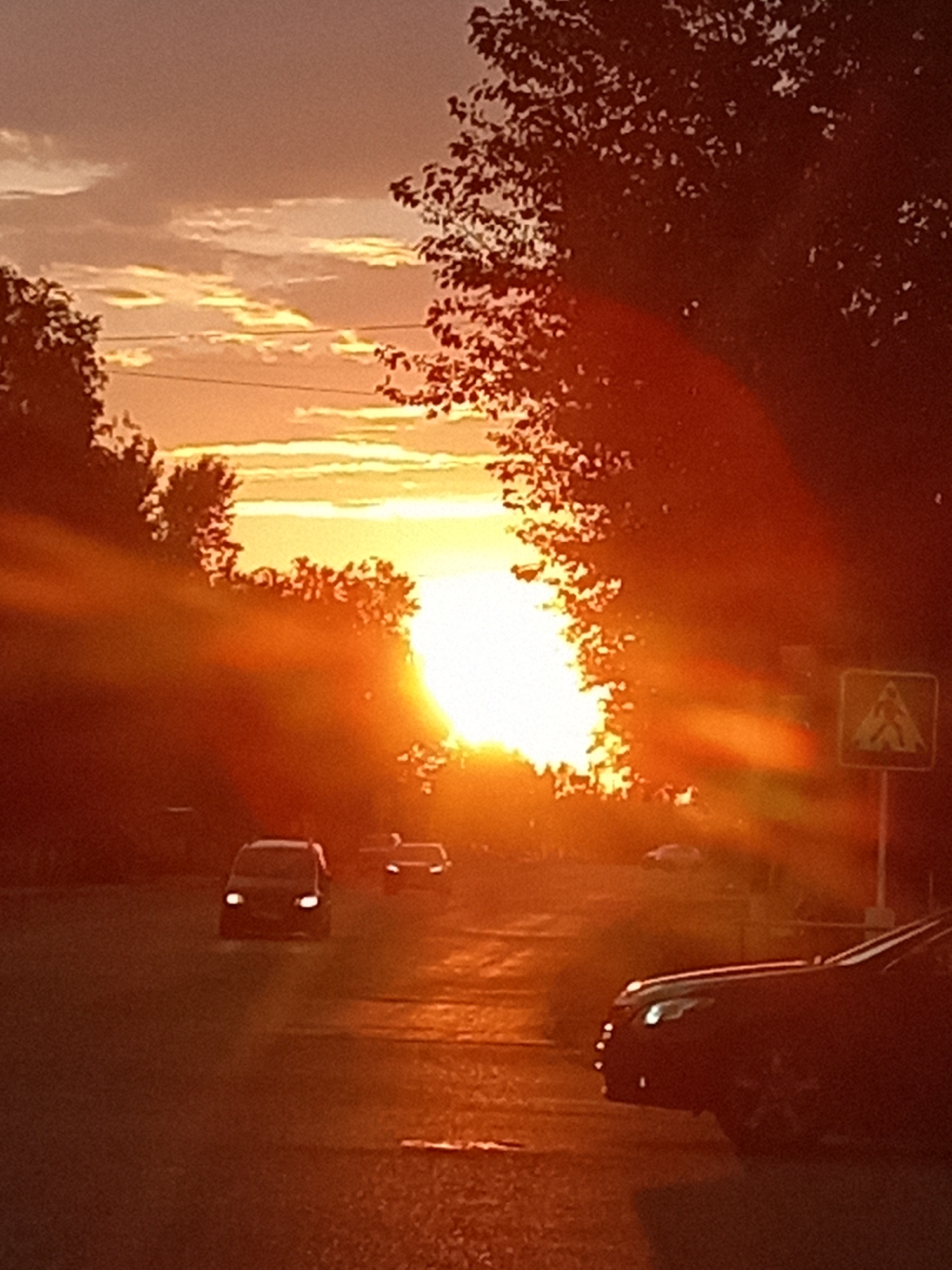 The image depicts a street scene in Rudny, Kazakhstan, during sunset. The sun is low on the horizon, casting a warm, orange glow across the scene and creating lens flare effects. A street is visible with several cars. In the foreground, a dark-colored car is parked on the right side of the road. Further down the street, a darker vehicle with its headlights on is driving away from the viewer. Another lighter-colored vehicle is also visible in the distance on the left side of the road, also with its headlights illuminated. Trees line the sides of the street, and their silhouettes are visible against the bright sky. A pedestrian crossing sign is present on the right side of the road. The sky is filled with clouds, illuminated by the setting sun. The overall impression is that of an evening commute or a moment of tranquility as the day ends.