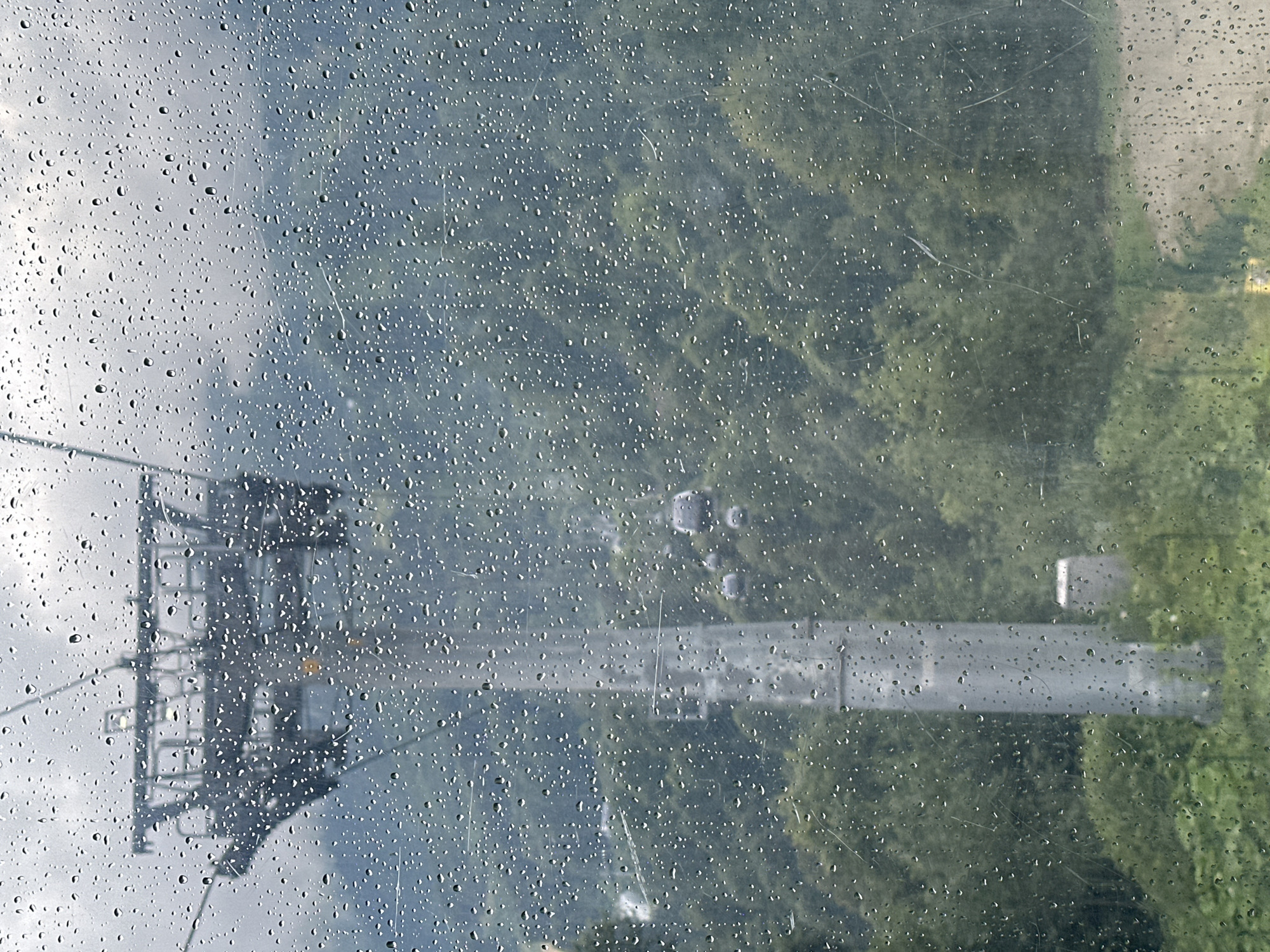 The image depicts a view through a rain-streaked window, likely from a cable car in Estosadok, Russia. In the foreground, numerous water droplets distort the view of the landscape. Dominating the left side of the frame is a tall, grey pylon for a ski lift or cable car system. It is a substantial concrete structure with visible supports and bracing at the top, where it anchors the cables. In the middle ground, several cable car cabins, appearing dark and rounded, are suspended from the cables, moving away from the viewer. The background is a dense, green forest that covers the slopes of mountains. The sky is overcast and grey, contributing to the overall moody atmosphere. The visual effect of the rain on the window creates a blurred and textured appearance, obscuring finer details of the distant scenery.