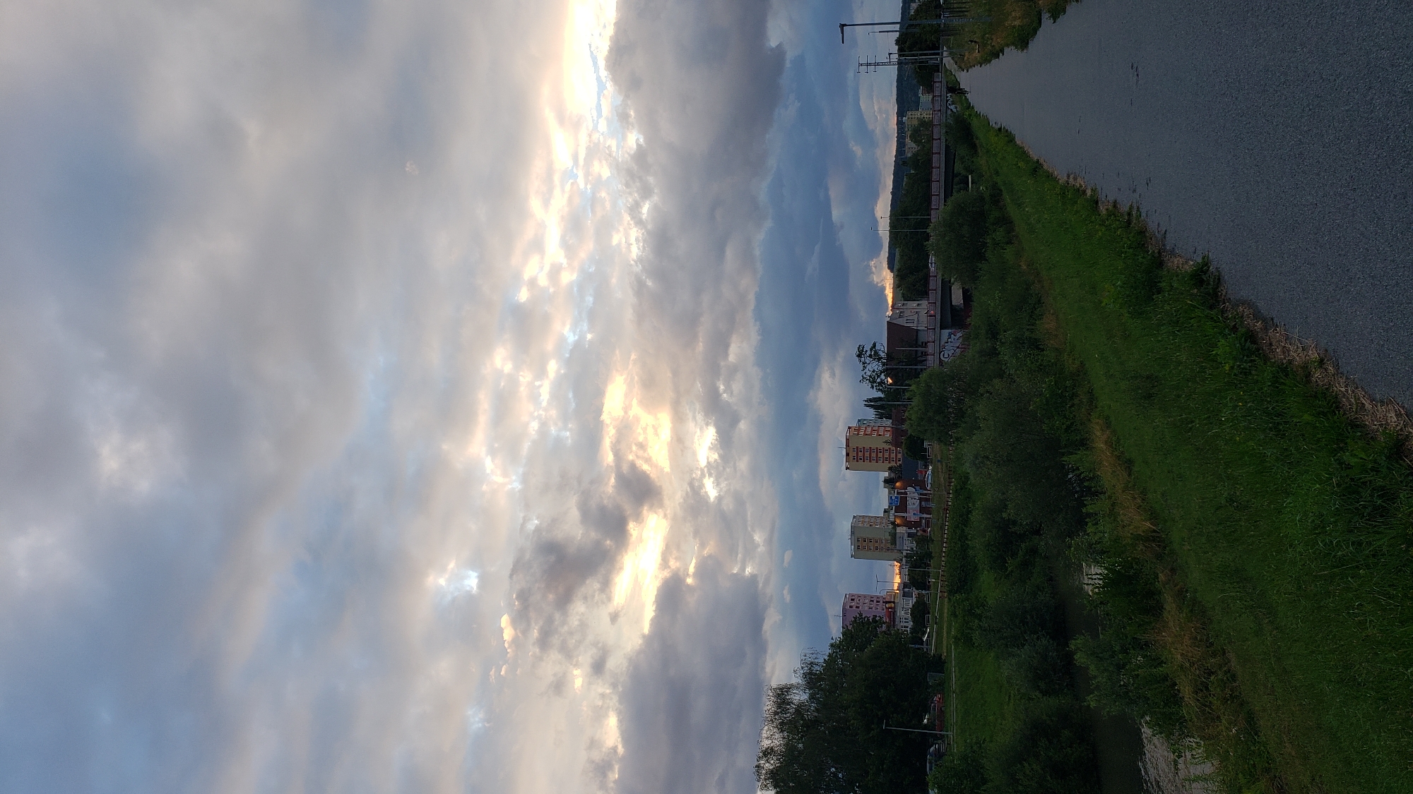 The image depicts a paved pathway alongside a grassy embankment and a body of water, likely the Torysa River, in Prešov, Slovakia. In the distance, three tall, multi-story buildings are visible, characteristic of the Sekčov housing estate. The sky is overcast with dramatic clouds, with patches of sunlight breaking through, suggesting either dawn or dusk. A bridge crosses the water in the mid-ground, and further along the pathway, a lone cyclist is visible. The overall scene conveys a sense of urban landscape merging with natural elements.