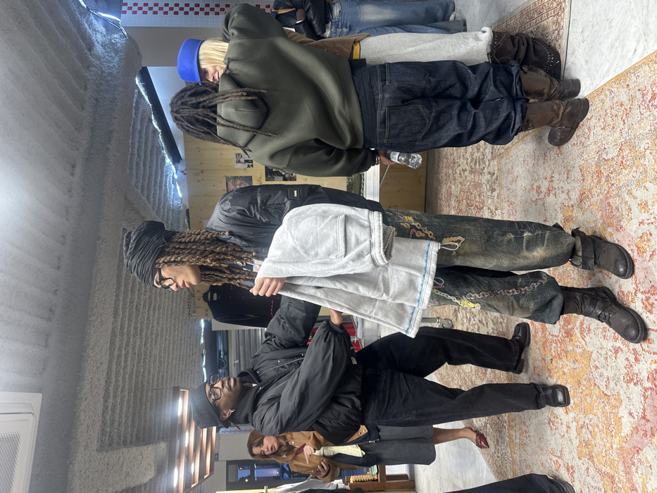 The image depicts an indoor scene within a commercial establishment, likely a boutique or gallery, in Paris, France, as indicated by a visible "Five Guys" logo in the upper right background.
In the foreground, a male with dark skin, wearing a black beanie, clear-framed glasses, a black puffer jacket, and black pants, stands with his right arm extended towards another individual. This central figure, also male, wears a black turban over long dreadlocks, clear-framed glasses, a black bomber jacket, and heavily embellished, distressed dark wash jeans featuring multiple chains. He holds a pair of light grey sweatpants in his left hand. The two appear to be engaged in an interaction or conversation.
To their right, another male, viewed from behind, wears dreadlocks, an olive green hooded sweatshirt, dark blue jeans, and brown boots. He holds a plastic water bottle. Standing closely beside him, facing partially away from the camera, is a female with blonde hair under a blue cylindrical hat, wearing a brown jacket and a light grey skirt. Further to the left in the mid-ground, a female with light brown hair, wearing a brown jacket and grey skirt with red shoes, holds a mobile phone.
The environment features a rough, textured grey ceiling, possibly with exposed elements and blue LED strip lighting. The floor is covered by a large, orange, red, and cream patterned rug. Background elements include wooden wall paneling, white walls, and what appears to be clothing displayed on racks or hangers.