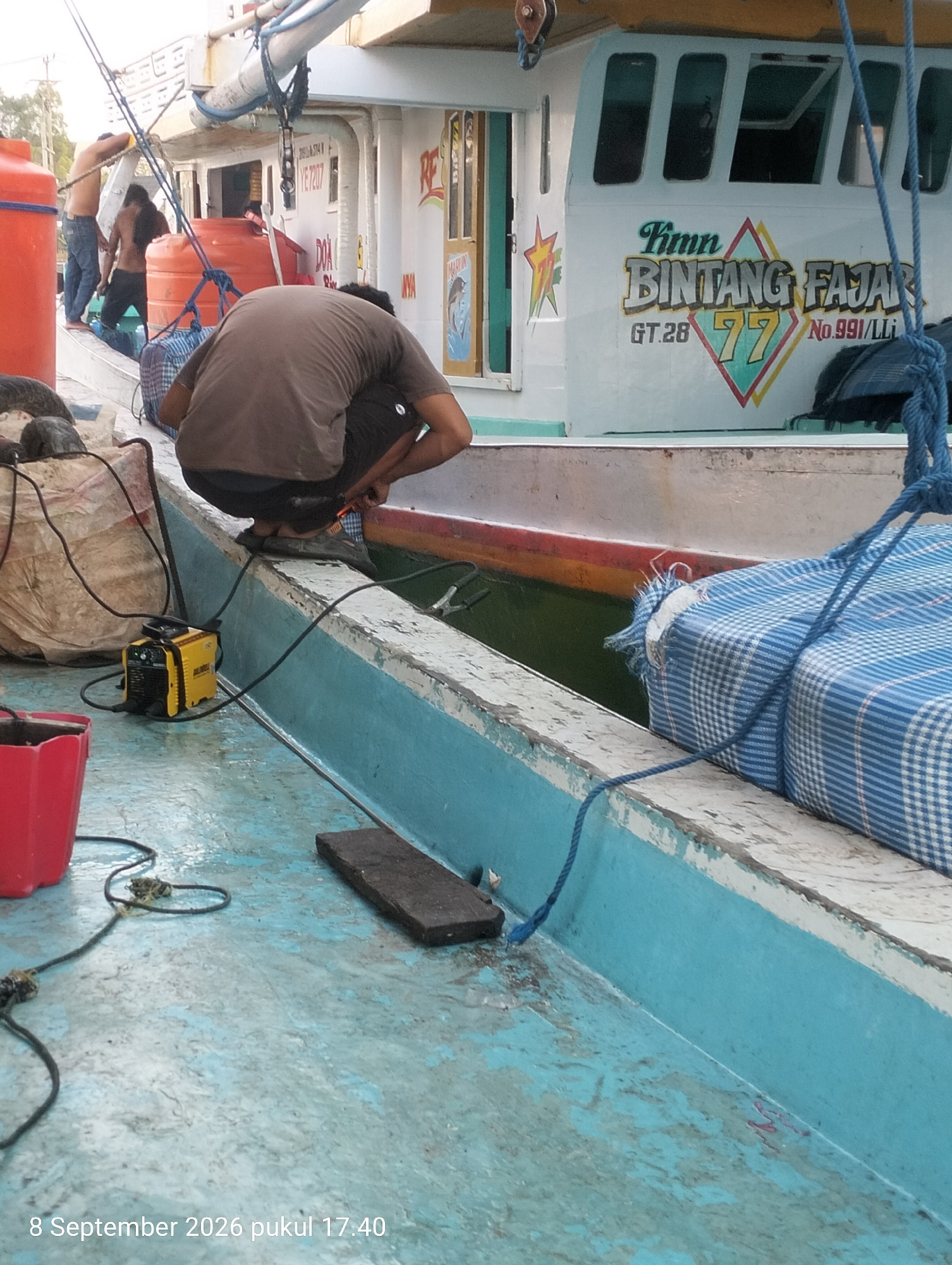 In Kendari, Indonesia, a person in a brown t-shirt and dark shorts is crouching on the deck of a boat, attending to some equipment. The boat, named "KM. BINTANG FAJAR" and identified as GT.28 No. 991/LLi, is white with blue accents along the edge of the deck. Other individuals are visible in the background, some shirtless, near large orange containers and bundled objects. A yellow welding machine is positioned on the deck near the crouching person, connected by cables to other parts of the boat. The deck itself is weathered and appears to be painted blue and grey, with some areas showing wear and tear. There are ropes, a red bucket, and a black rectangular object also present on the deck. The water visible beneath the deck is green. The timestamp "8 September 2026 pukul 17.40" is overlaid on the bottom left of the image.