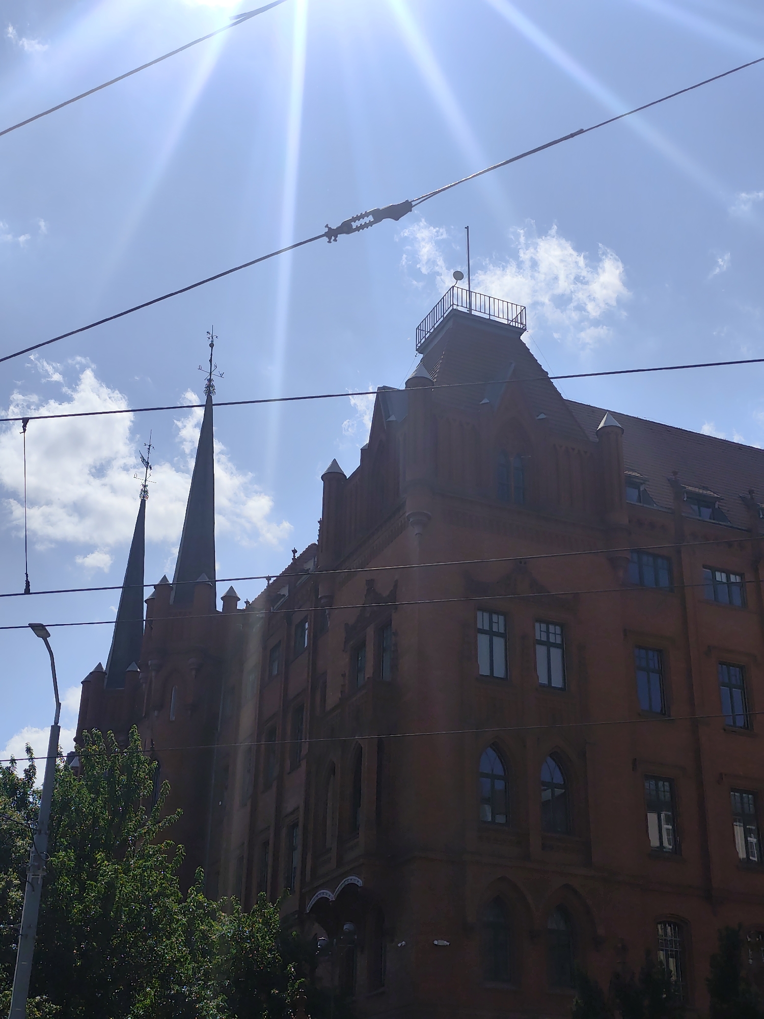 The image depicts the historic red brick building of the Old Town Hall (Ratusz Staromiejski) in Szczecin, Poland. The building features distinctive Gothic architecture with pointed spires and ornate detailing. Several wires, likely from a tram or trolleybus line, crisscross the foreground, partially obscuring the view of the structure. The sun is shining brightly, casting a lens flare across the sky, and some clouds are visible. Lush green trees can be seen at the base of the building on the left side of the frame. The perspective is from a low angle, looking upwards at the building.