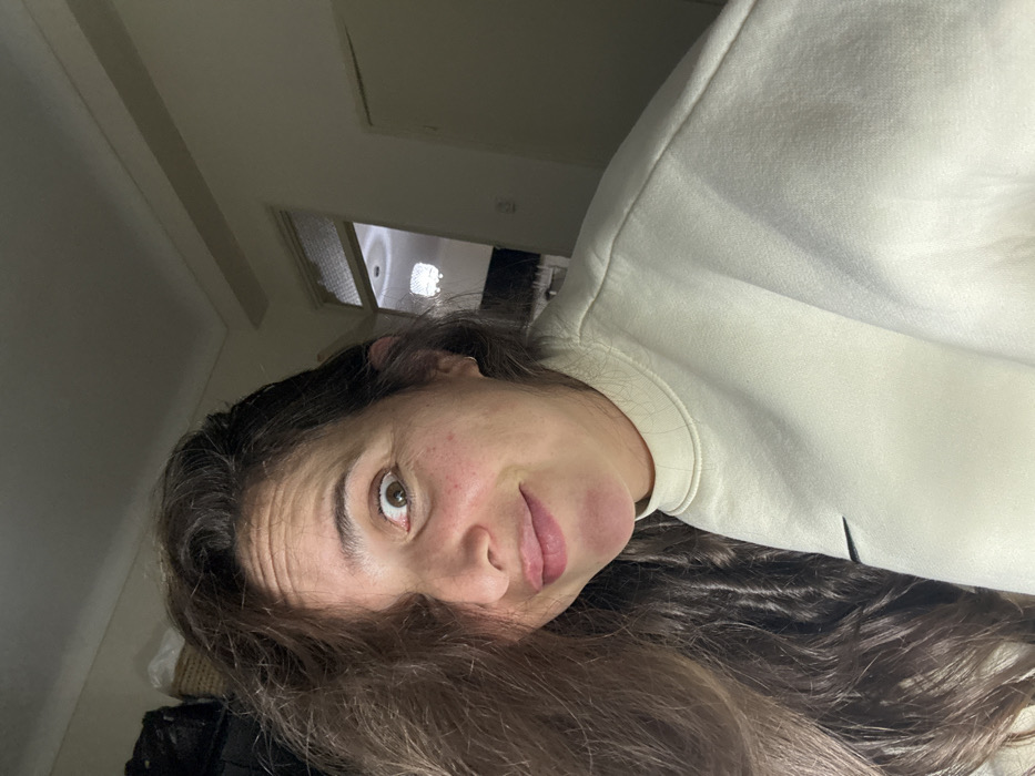 A woman with brown hair, wearing a cream-colored sweatshirt, poses for a selfie. She is indoors, and the lighting is subdued. She has brown eyes and fair skin with some visible freckles. The background shows a glimpse of a room with a white ceiling and a doorway. Her head is tilted slightly, giving a casual and intimate feel to the image.