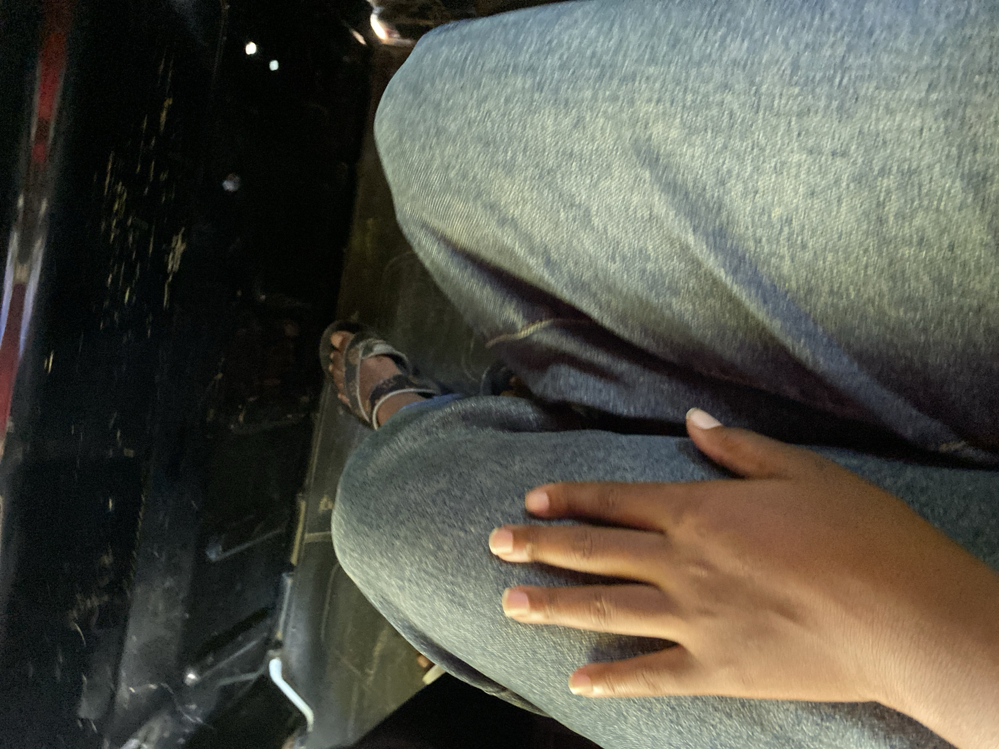 The image is a high-angle shot of a person sitting, wearing denim jeans and brown sandals. The person's hand rests on their leg. To the left, there is a dark, possibly textured surface, which could be part of a car interior. No specific details suggest it is in Dar es-Salaam, Tanzania.
