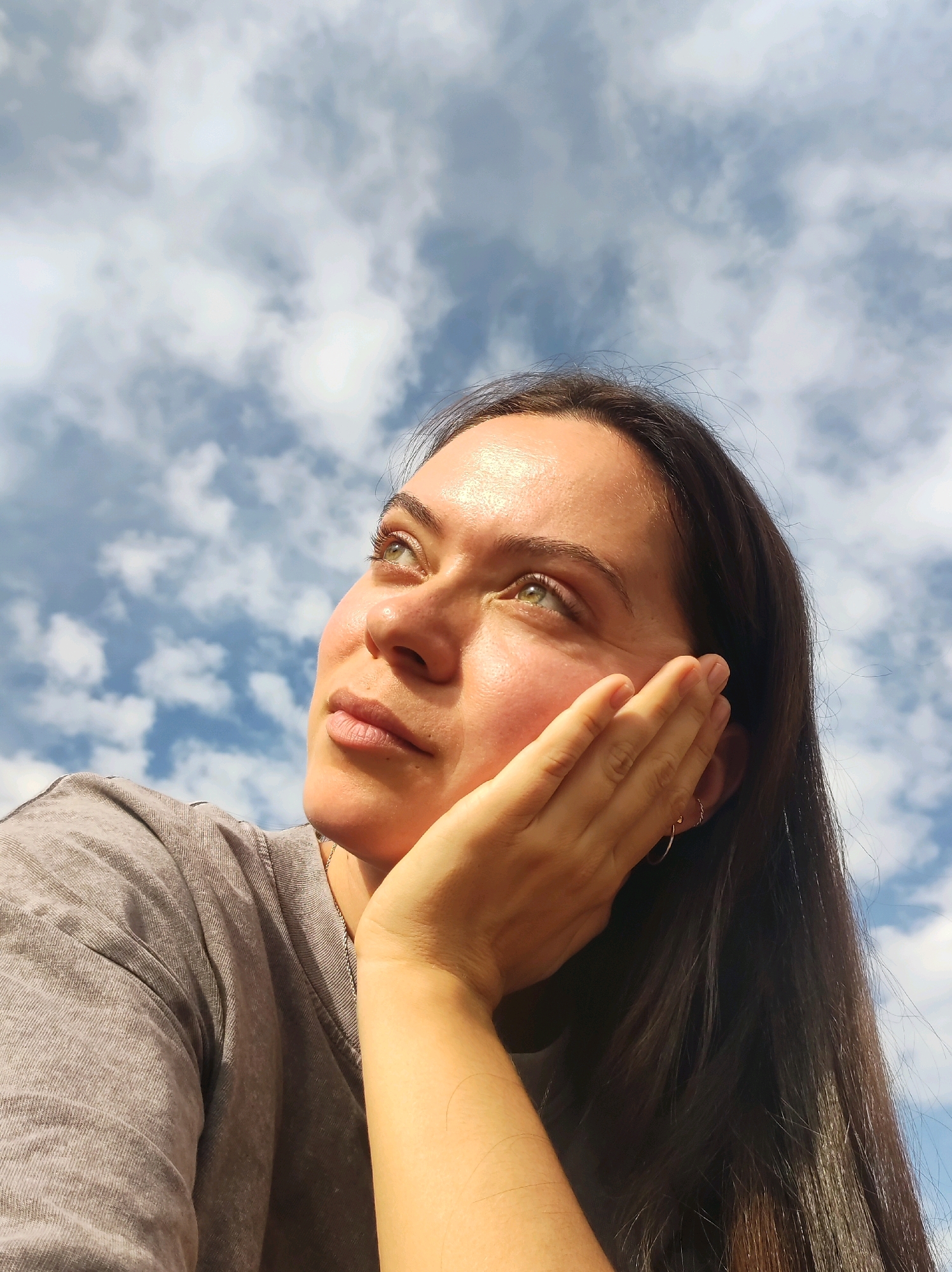 The image shows a close-up, waist-up shot of a young woman with long, dark hair, looking upwards and to her left. She is wearing a grey t-shirt. Her right hand is raised to her face, resting on her cheek and chin, with her fingers gently touching her face. She has green eyes, and her skin appears to be illuminated by sunlight. Her expression is one of contemplation or wonder. The background consists of a blue sky with scattered white clouds, suggesting an outdoor setting on a bright day. The image is taken from a low angle, looking up at the woman and the sky. There is no identifiable location within the city of Chaikovsky, Russia, depicted in this image.