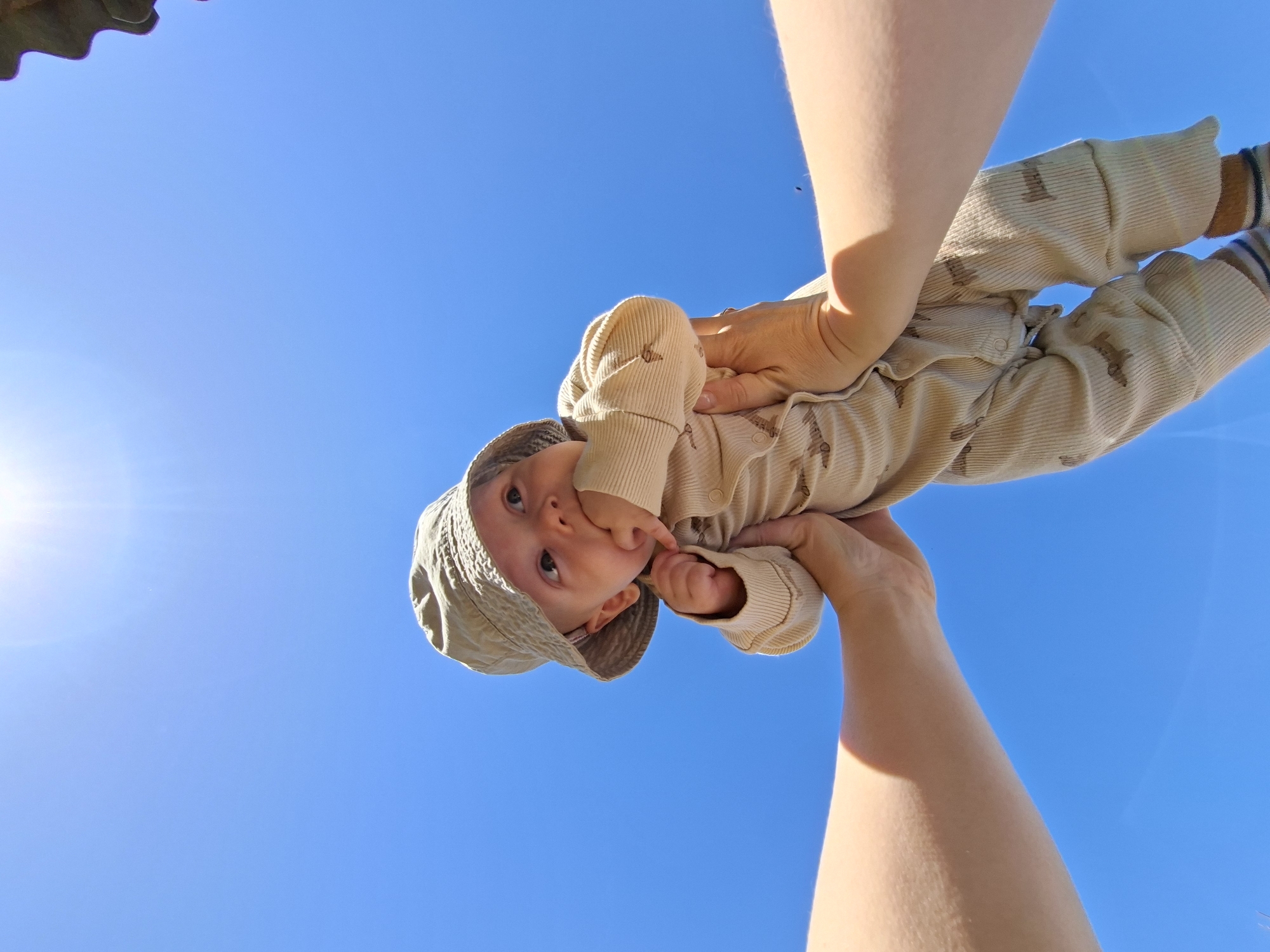 A low-angle photograph shows a baby being held up against a clear, blue sky. The baby wears a light-colored outfit and a hat. The baby's fingers are near its mouth. Two fair-skinned adult arms hold the baby. The top-left corner of the image shows what appears to be a roofing detail. Karsha, Russia, is not explicitly indicated.