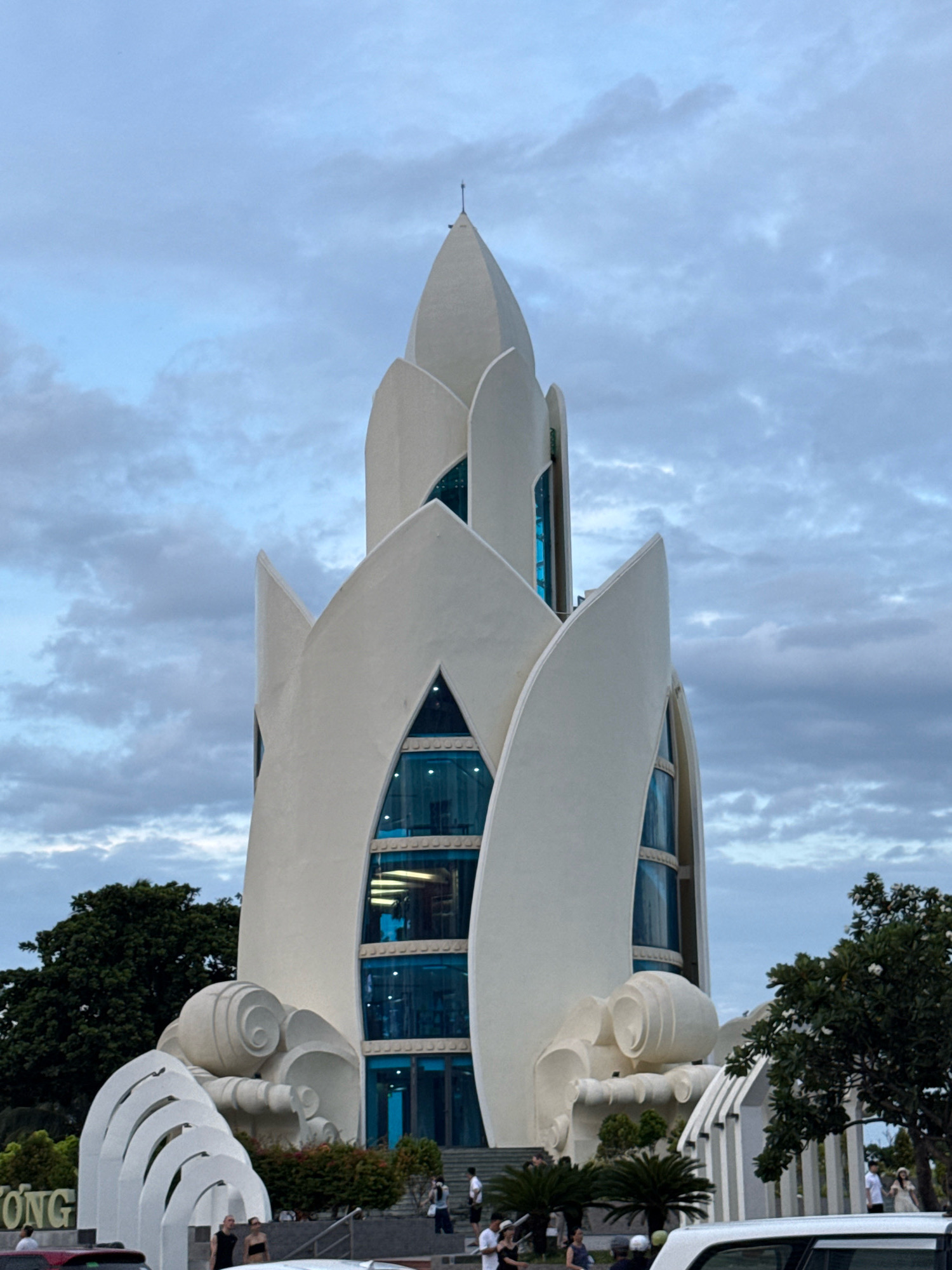 A tall, white, modern building with a petal-like design resembling a lotus flower dominates the center of the image. The building features large, triangular blue glass windows that offer glimpses of an interior with visible lighting. The structure is set against a cloudy, pale blue sky. In the foreground, a white arched structure with decorative swirls is visible, possibly a sculptural element or part of an entrance. Several people, dressed casually, are walking near the building and on stairs leading up to it. Trees, including a large dark green tree on the left and a smaller tree with white flowers on the right, frame the scene. The bottom edge of the image shows the tops of vehicles, indicating a public area or street. The building is identified as the Quan Am statue at Long Son Pagoda in Nha Trang, Vietnam.