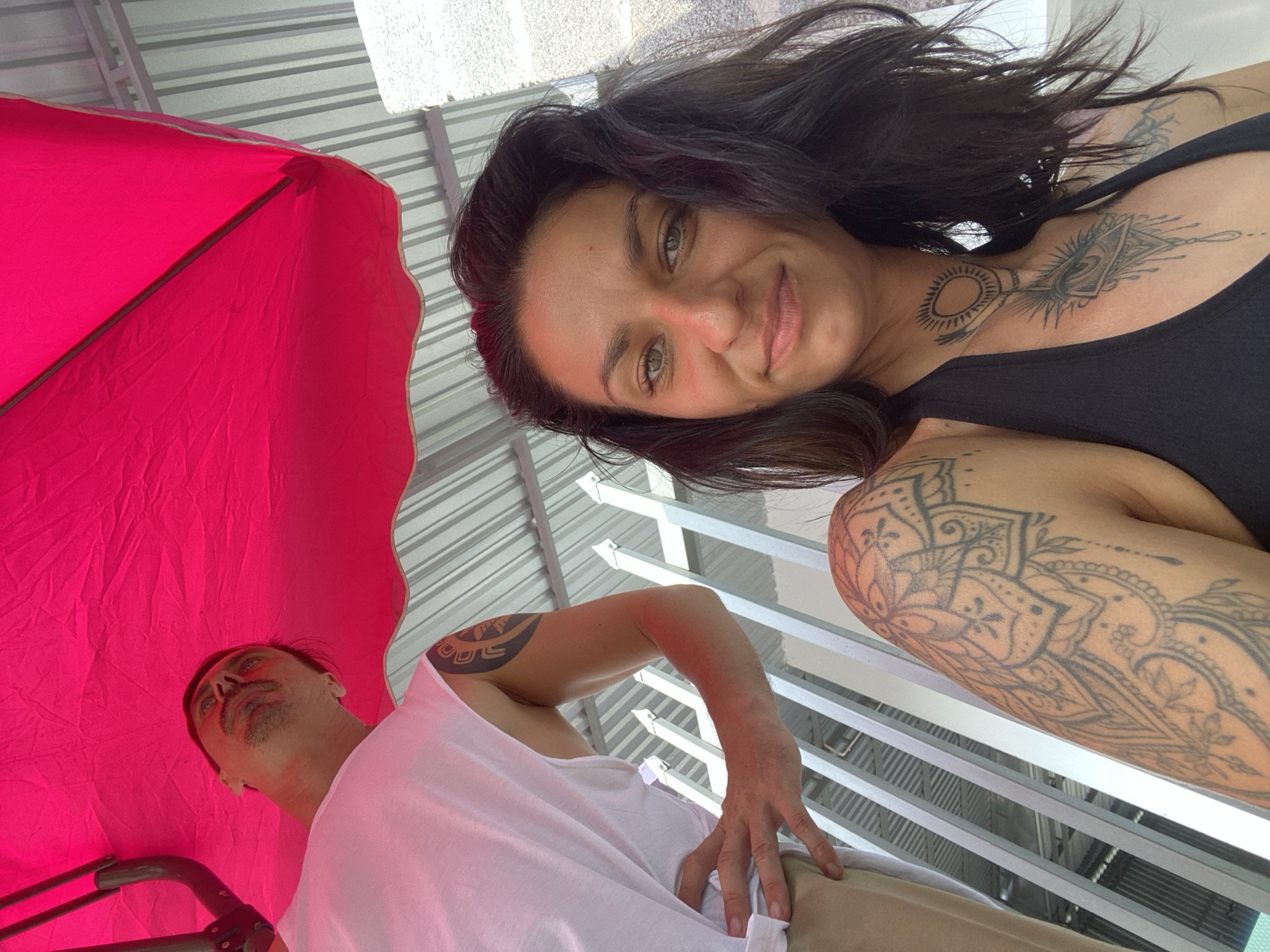 An outdoor, waist-up, medium shot shows two people in Mũi Né, Vietnam, posing under a bright pink umbrella. The woman in the foreground, facing the camera, has dark, wavy hair, green eyes, and multiple tattoos, including a large mandala on her left shoulder and chest. She is wearing a black top. Behind her and to the left, a man in a white tank top looks upwards and away from the camera. He has a tattoo on his left arm and stubble on his face. The background consists of a corrugated metal roof and a white metal fence. The lighting suggests a sunny day.