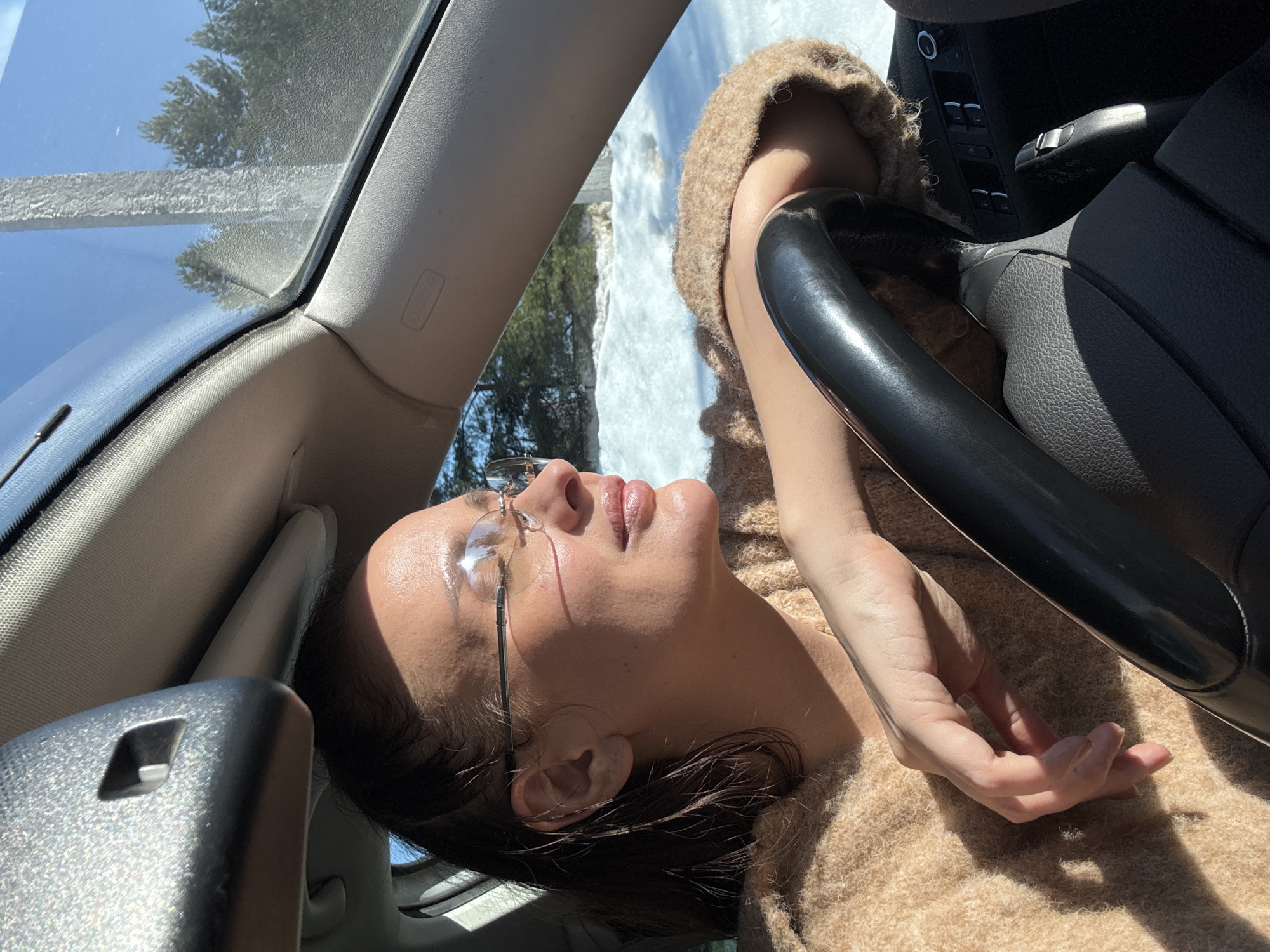 A woman with dark hair and wearing eyeglasses lies in the driver's seat of a car, her head tilted back against the headrest. She is wearing a tan sweater. Her right arm rests on the steering wheel. The interior of the car is visible, including the dashboard and window controls. Outside the car, a snowy landscape with trees is visible through the windshield. The location is inferred as Chaikovsky, Russia.