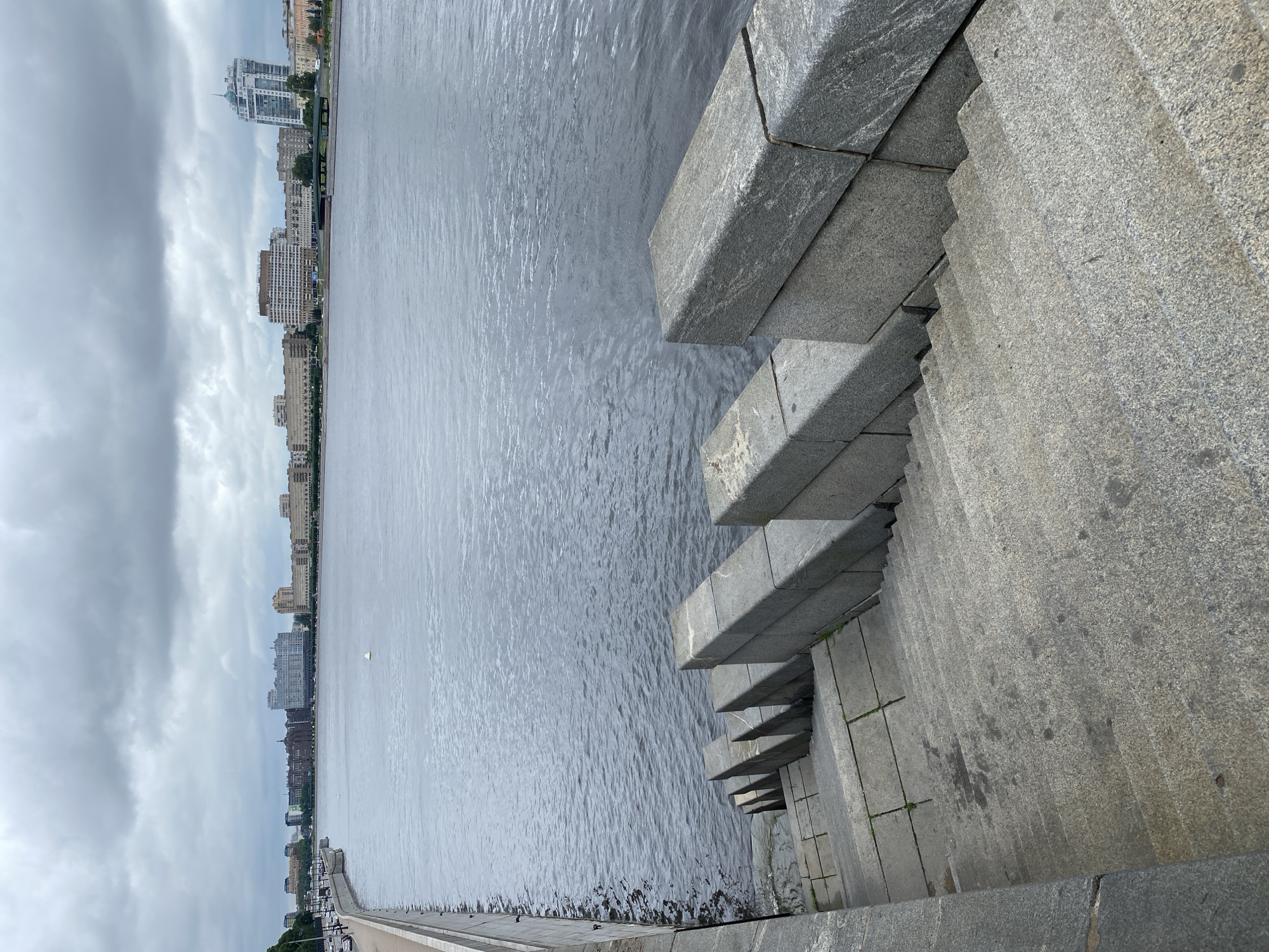 A body of water, likely a river, occupies the majority of the frame. The water's surface is rippled with a moderate chop, reflecting the overcast sky above. The sky is filled with dense, gray clouds, suggesting a cloudy or potentially rainy day.
In the foreground, a wide set of granite stairs descends towards the water. The stairs are constructed from large, rough-hewn blocks of gray stone, showing signs of wear and some moisture. The steps are segmented by horizontal granite slabs, with small patches of grass growing between some of them. The sides of the stairs are lined with similarly constructed granite walls.
Across the water, a cityscape is visible. A long stretch of buildings lines the distant shore, varying in height and architectural style. These buildings appear to be a mix of residential and commercial structures, with some taller, modern buildings interspersed with older, more traditional designs. A bridge is faintly visible in the mid-ground, spanning the water. The left edge of the frame shows a paved embankment along the water's edge, with a few cars parked on it.