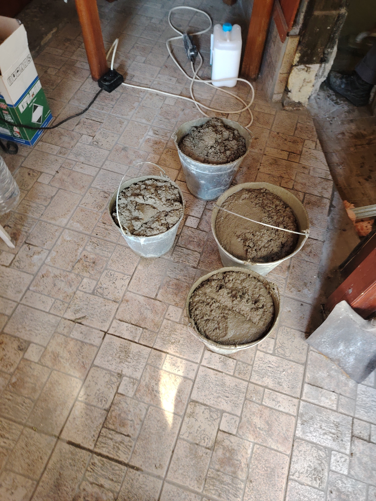 The image shows an indoor scene on a tiled floor. Four buckets, appearing to contain a mixture resembling wet cement or mortar, are arranged in a somewhat circular pattern. To the top left is a partially visible cardboard box with green accents. Cables and cords are scattered near the top center, accompanied by a white plastic container. A section of a table leg is visible in the background. To the far right, a person's leg and foot, clad in a dark shoe, are slightly visible. The surrounding environment suggests a room undergoing renovation or construction. While the image provides details about the interior, it does not contain information indicating the location in Panagyurishte, Bulgaria.
