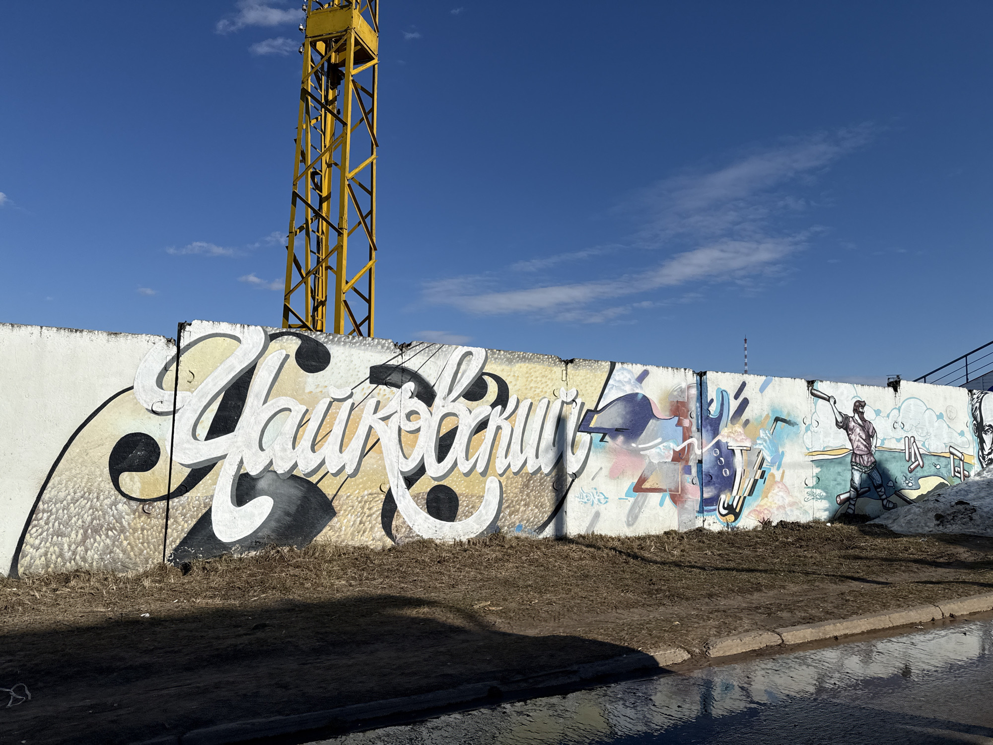 The image shows a section of a concrete wall adorned with graffiti. The wall is in the city of Unknown, Unknown. Prominent in the image is the graffiti featuring the word "Чайковский" (Tchaikovsky) in large, stylized white Cyrillic letters with black accents. The rest of the graffiti comprises colorful abstract and figurative elements, including a depiction of a person.
Behind the wall, there is a yellow construction crane extending upward. The ground in front of the wall has a mix of dry grass and patches of melting snow or damp earth. A wet street is visible in the foreground. The sky is bright blue with scattered clouds.