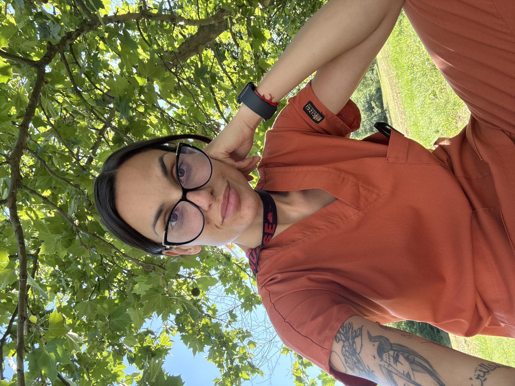 A young woman with dark hair and glasses is sitting outdoors under a tree, her head resting on her hand. She is wearing an orange scrub top and pants. A black choker with red lettering is around her neck. She has tattoos on her left arm and a watch on her right wrist. A pen is visible in the pocket of her scrub top. The background consists of green grass and trees. This image was taken in Pleven, Bulgaria.