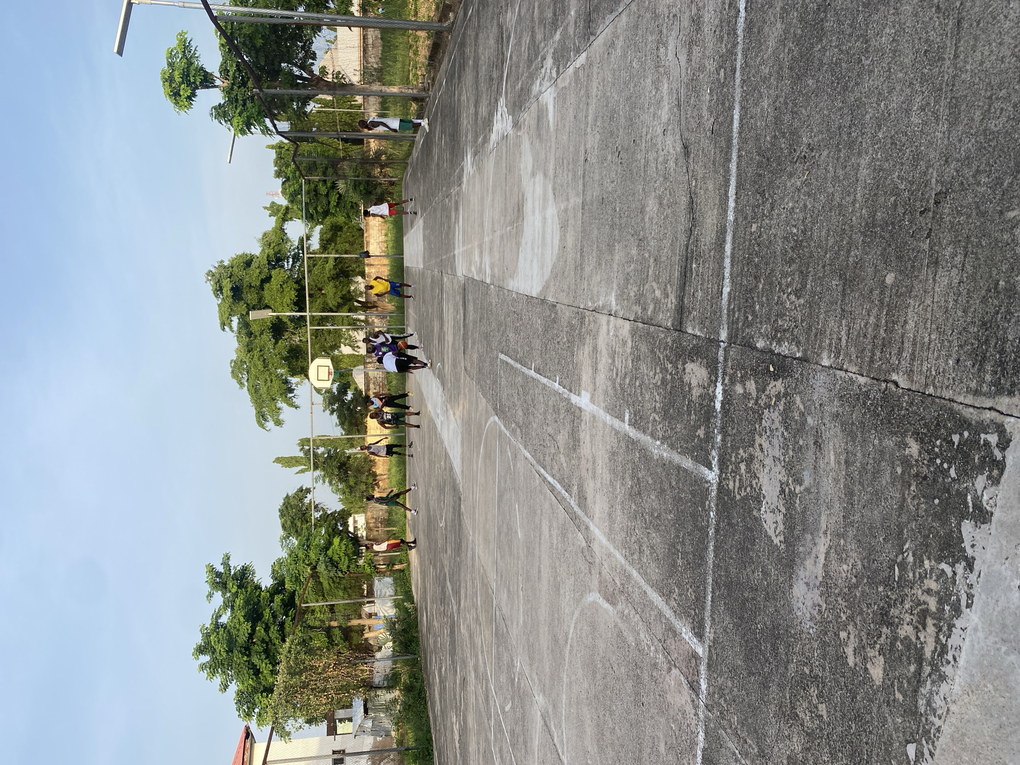 A group of young males are playing soccer on a concrete court in Jalingo, Nigeria. The court is marked with white lines. A basketball hoop is visible in the background. Several trees and a fence surround the court. The sky is clear and blue. The people are wearing athletic clothing.