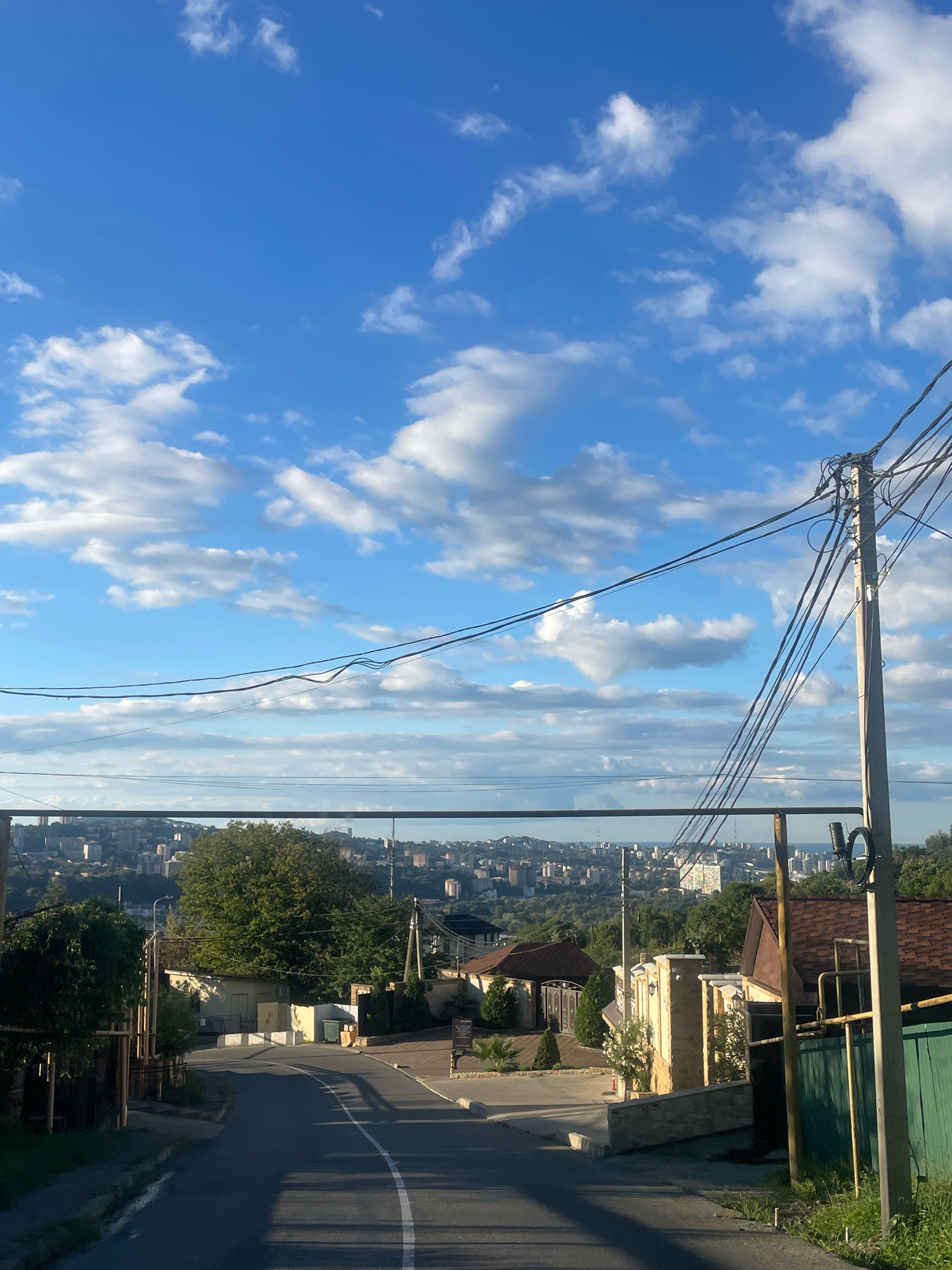 The image depicts a street scene in Baranovka, Russia, on a partly cloudy day. A paved road curves to the left, leading downhill, with a white dashed line marking the center. On the right side of the road, there is a utility pole with multiple wires extending across the frame. Adjacent to the pole is a green fence and a house with a brown, shingled roof and a light-colored stone wall. Further down the street, on the left, are trees and fences. In the background, a cityscape with buildings and trees stretches across the horizon under a blue sky dotted with white clouds. The lighting suggests it is daytime. There are no people or animals visible in the image.