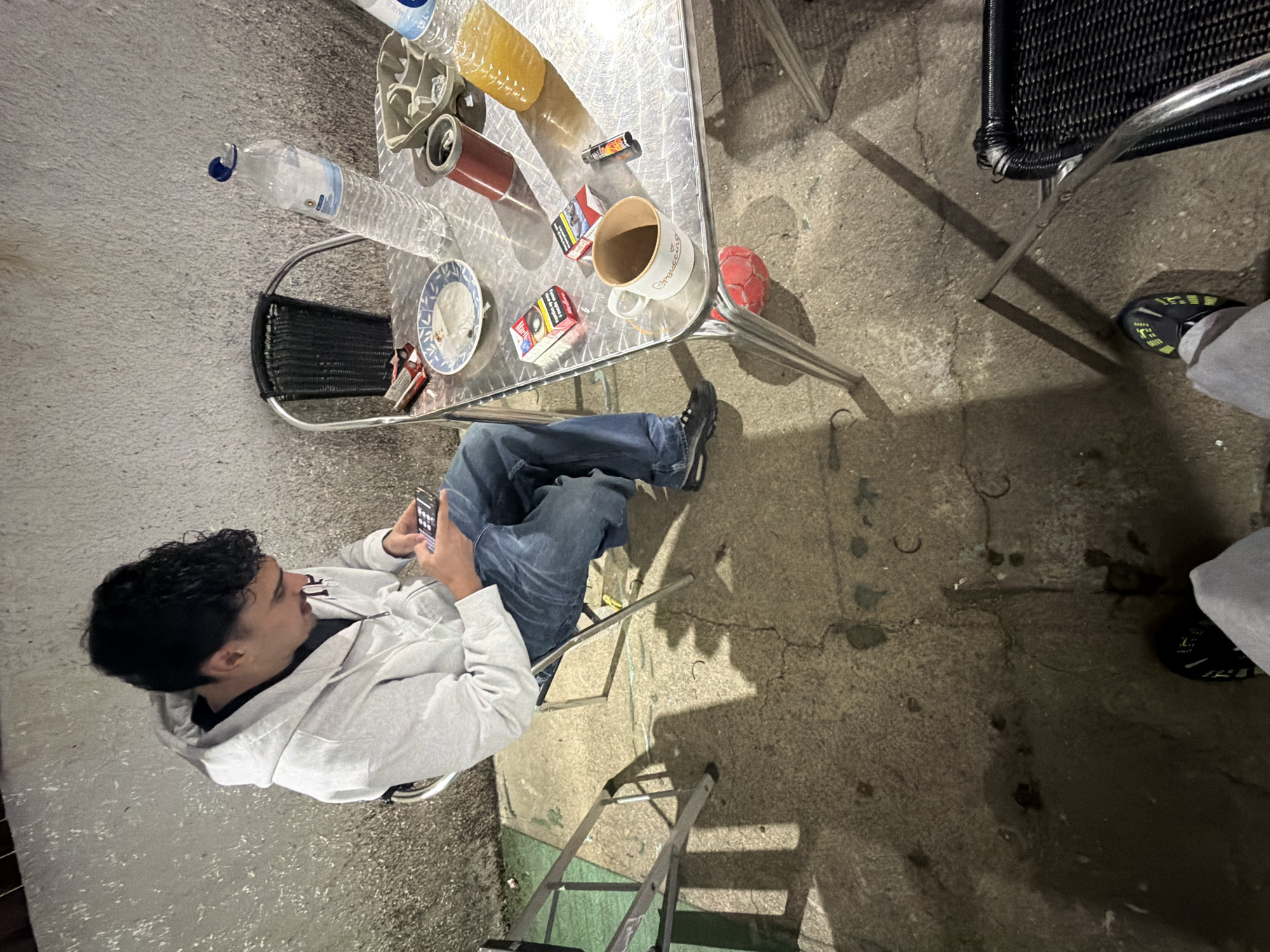 A person wearing a grey hoodie and blue jeans sits in a chair, looking at a mobile phone. The scene is outdoors, possibly a patio or backyard, given the concrete flooring and adjacent wall. A metal table is cluttered with various items, including a water bottle, a bottle of orange liquid, a coffee mug, a red ball, cigarettes, and a lighter. Another chair is visible nearby. A ladder is placed on the ground adjacent to the sitting person. The photograph provides no information to determine the location in Vila Real, Portugal.