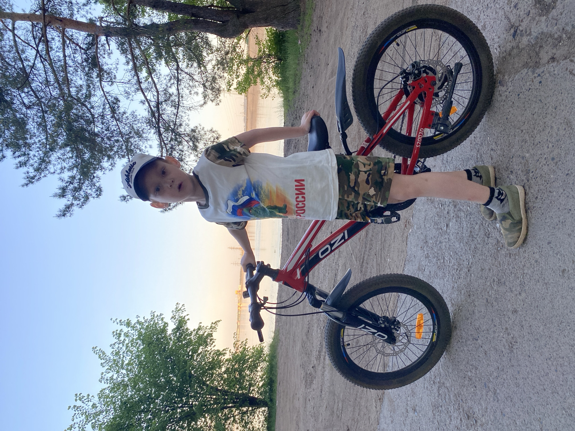 A child in camouflage shorts, an olive green shirt, and a hat stands beside a red and black OZI brand bicycle. The child is looking at the camera. The child is wearing olive green shoes and socks with a pattern on them. The backdrop shows a tree and possibly a body of water in the distance, and there's ground beneath. The shirt has a print of the Russian flag and says "РОССИИ" in red. This appears to be an outdoor setting in Noviy, Russia.