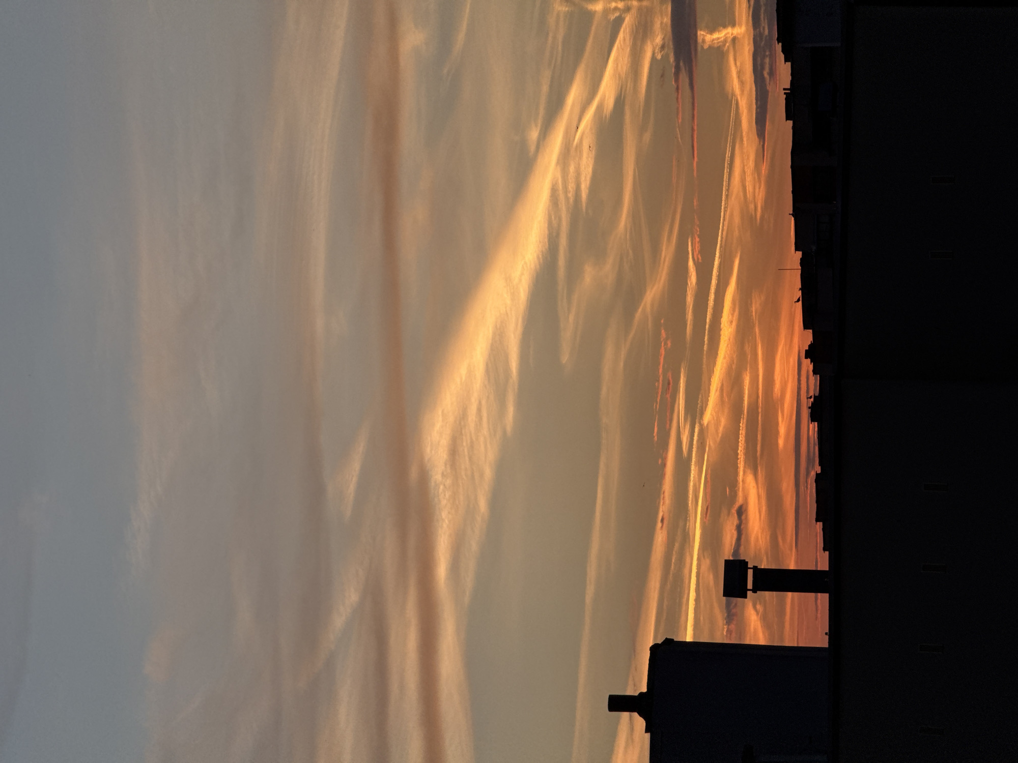 The image shows a sunset in Pleven, Bulgaria, with contrails streaking across the sky. The sky is mostly orange and yellow with a touch of blue. The silhouette of a building is visible on the right side of the frame.