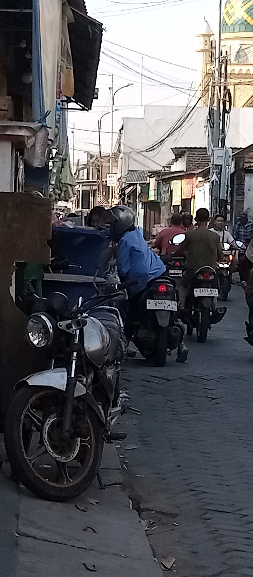 The image depicts a street scene in Wadungasri, Indonesia, characterized by a narrow, paved road flanked by buildings and a mosque visible in the background. Several motorcycles are present, some ridden by individuals wearing helmets. In the foreground, a silver and black motorcycle is parked on the left. Further down the street, a person in a blue shirt and helmet is seated on a motorcycle. Other individuals on motorcycles are also visible in the midground, with a mosque featuring a green and yellow dome and a minaret visible in the distance on the right. The environment appears to be an urban setting with overhead electrical wires and a somewhat cluttered streetscape, suggesting a busy, functional area.