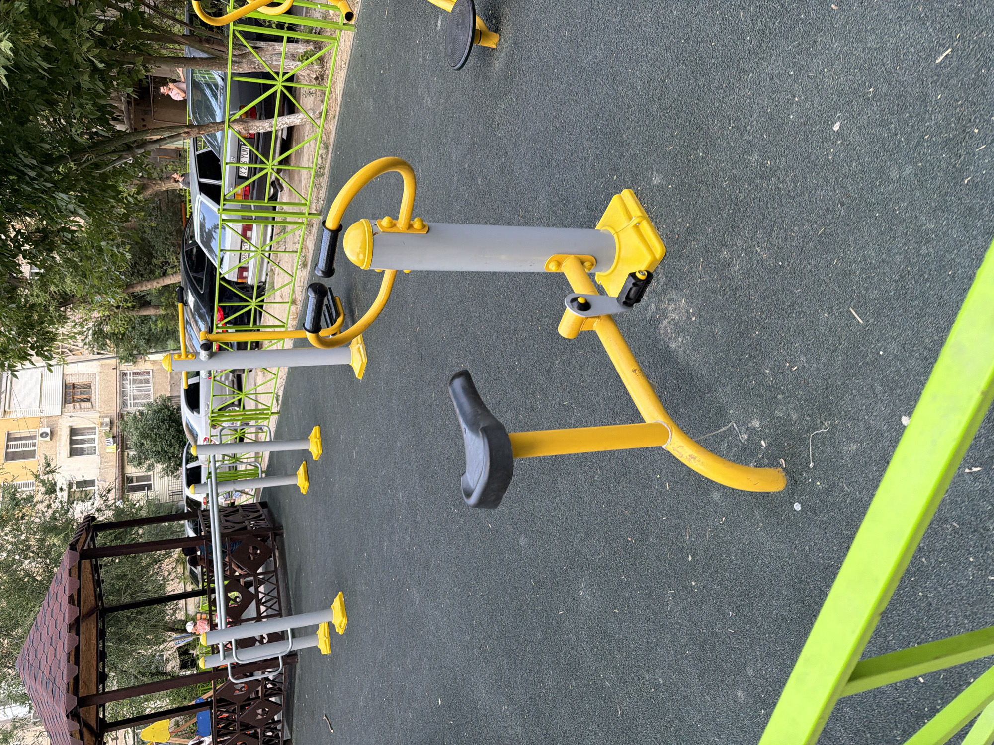 The image depicts an outdoor fitness area in Shymkent, Kazakhstan, situated within a residential complex, as indicated by the apartment buildings visible in the background. The foreground features several pieces of yellow and gray exercise equipment with black seats and pedals, designed for various physical activities. The ground is covered in a dark, textured surface, likely rubberized safety flooring common in playgrounds and outdoor fitness zones. A bright green railing runs along the bottom edge of the frame, suggesting a boundary or safety feature. In the middle ground, a brown gazebo-like structure with a red roof is present, and a green metal fence separates the fitness area from a street where several cars are parked. A few individuals are casually present, with one person standing near the fence and another sitting inside the gazebo, suggesting a relaxed, community atmosphere. The overall scene conveys a sense of public amenity and recreational space within an urban setting.