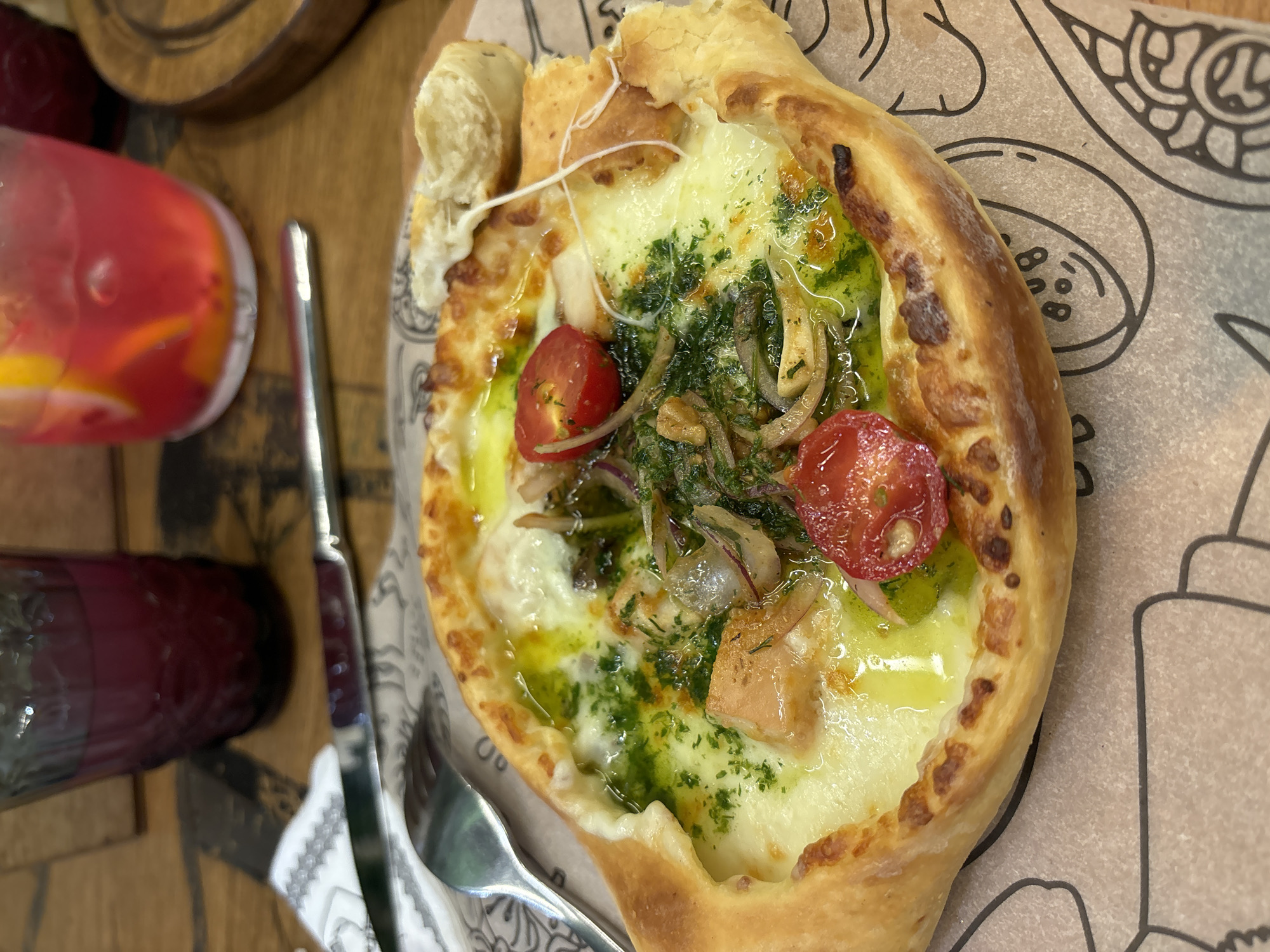 A close-up, eye-level shot shows a dish of khachapuri served on a wooden table with a decorative paper liner. The khachapuri, a boat-shaped bread filled with cheese and toppings, is golden brown with visible cheese pull. The toppings include halved cherry tomatoes, chopped herbs, thinly sliced red onions, and pieces of salmon. A fork and knife are visible to the left of the dish, and two glasses containing colorful drinks are in the background. The overall lighting is warm and inviting, suggesting an indoor setting. The scene is reminiscent of a meal at a restaurant in Izhevsk, Russia, where khachapuri is a popular and beloved dish.